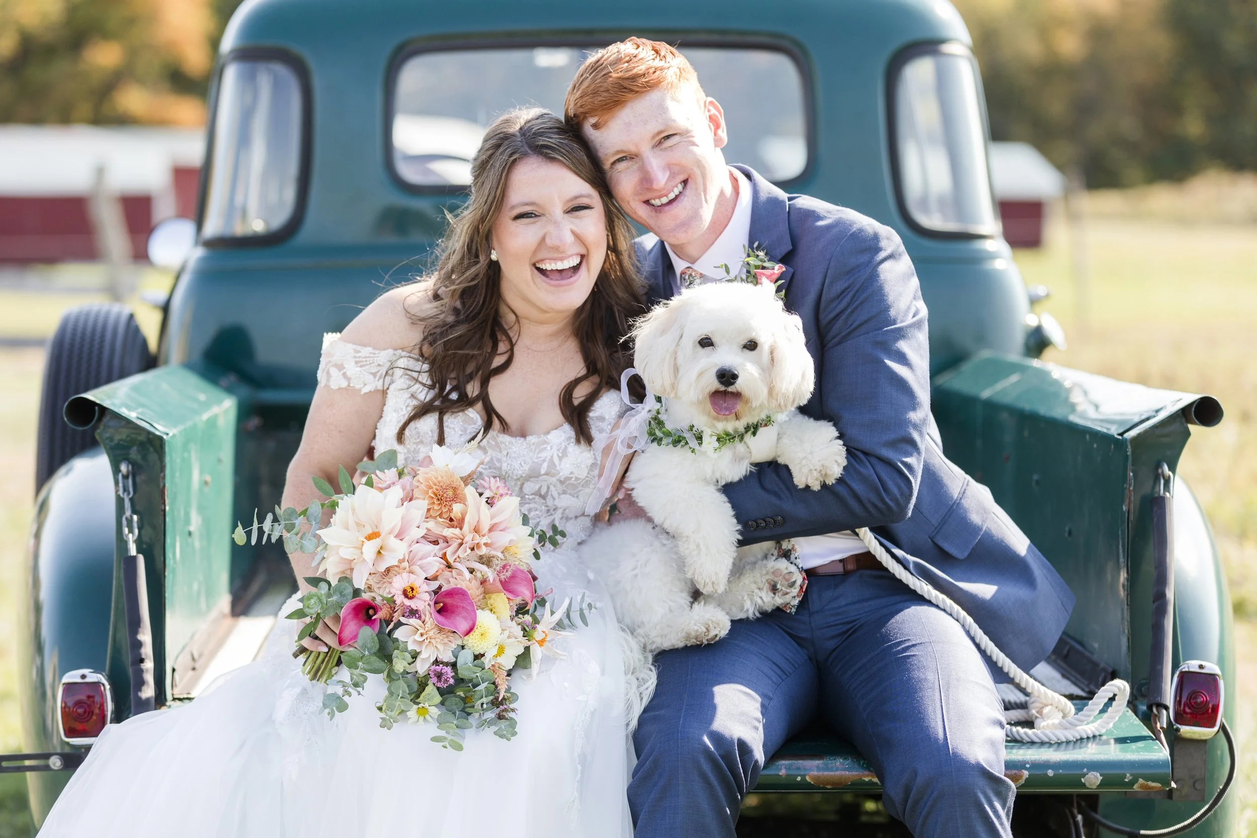 A newlywed couple sitting on the back of a green pickup truck, holding a white dog with a flower collar, smiling at the camera, with a bride holding a bouquet of flowers.