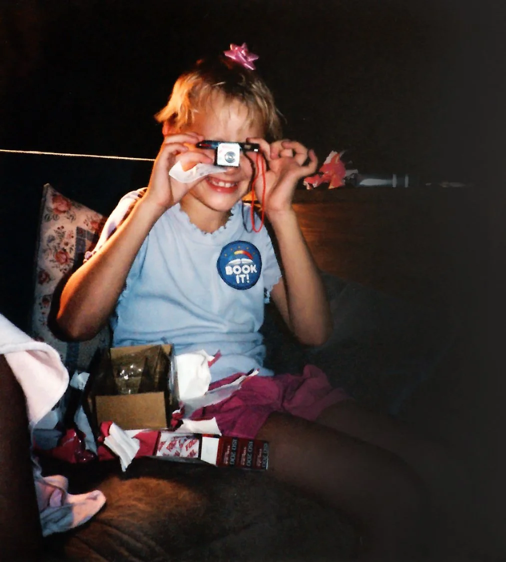 A young girl with blonde hair, sitting on a dark-colored couch, smiling and holding a small camera to her eye. She wears a white t-shirt with a sticker that says 'BOOK IT!' and pink shorts, with birthday decorations around her.