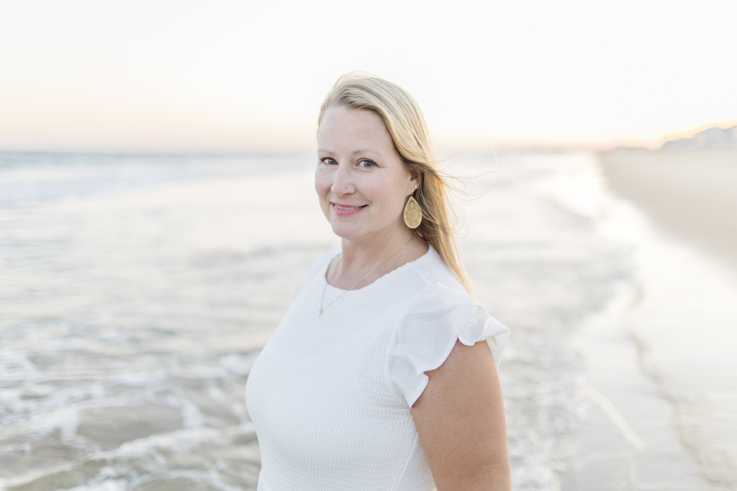 A woman with blonde hair smiling at the camera on a beach at sunset, wearing a white top with ruffled sleeves and gold earrings.