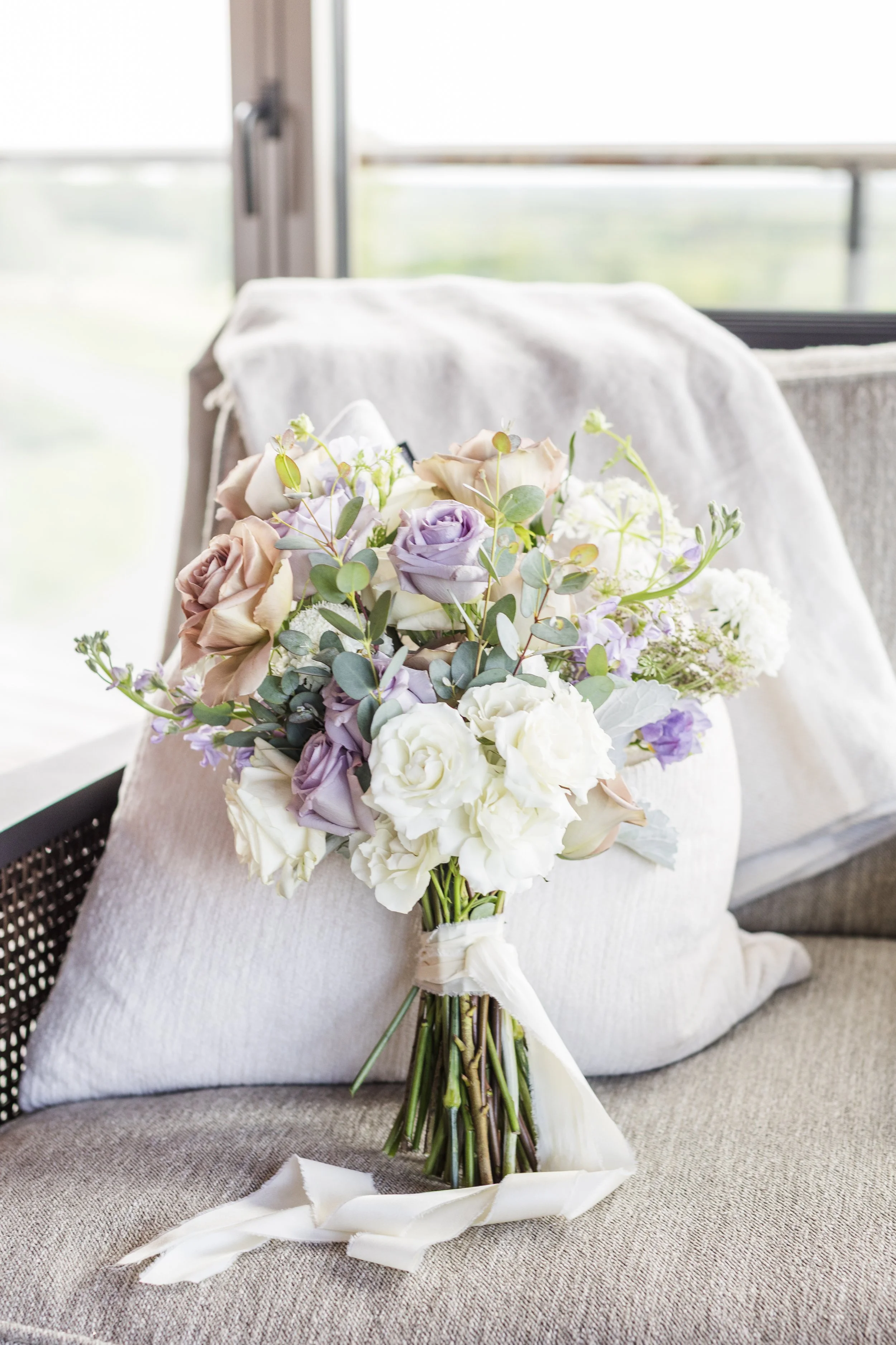 A bouquet of pastel-colored roses, lisianthus, and eucalyptus leaves on a beige sofa with pillows, in a bright room by a window.