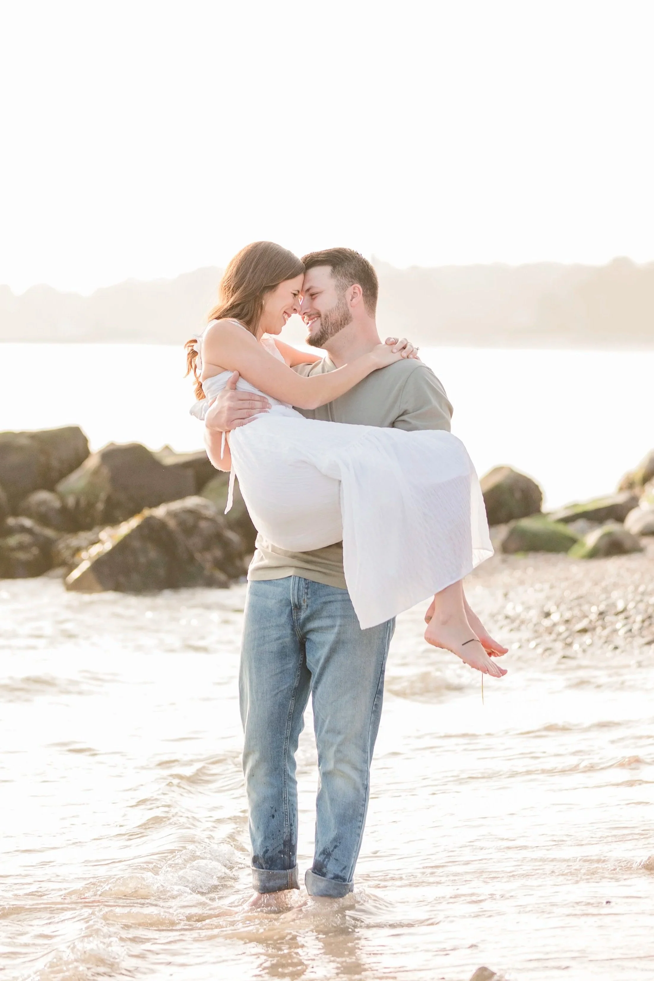 A man holding a woman in a white dress over shallow water on a beach, smiling and touching foreheads at sunset or sunrise.