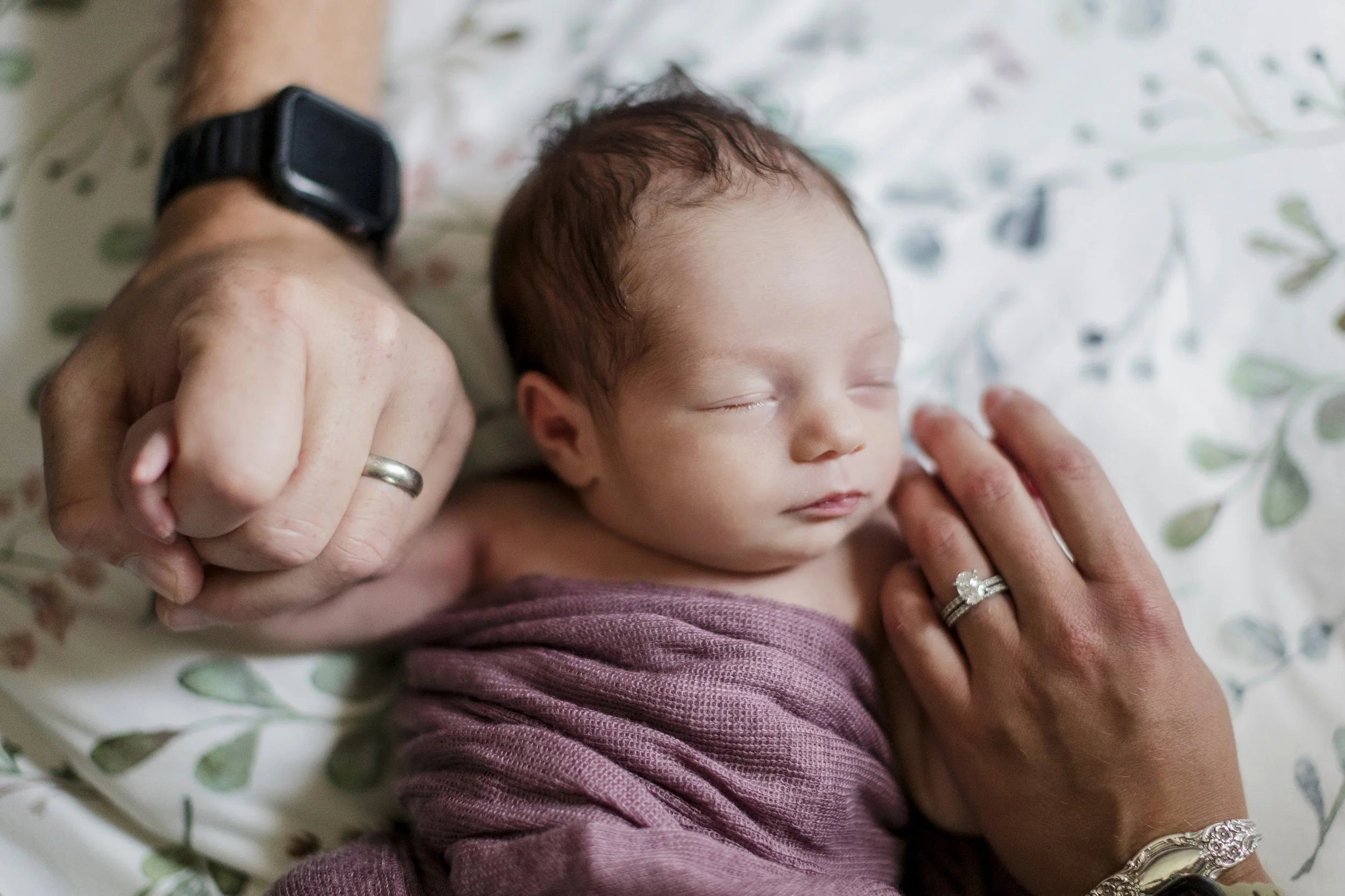 A newborn baby sleeping, wrapped in a purple blanket, being gently held by two adult hands with rings and a wristwatch. The baby has closed eyes and shows a peaceful expression.