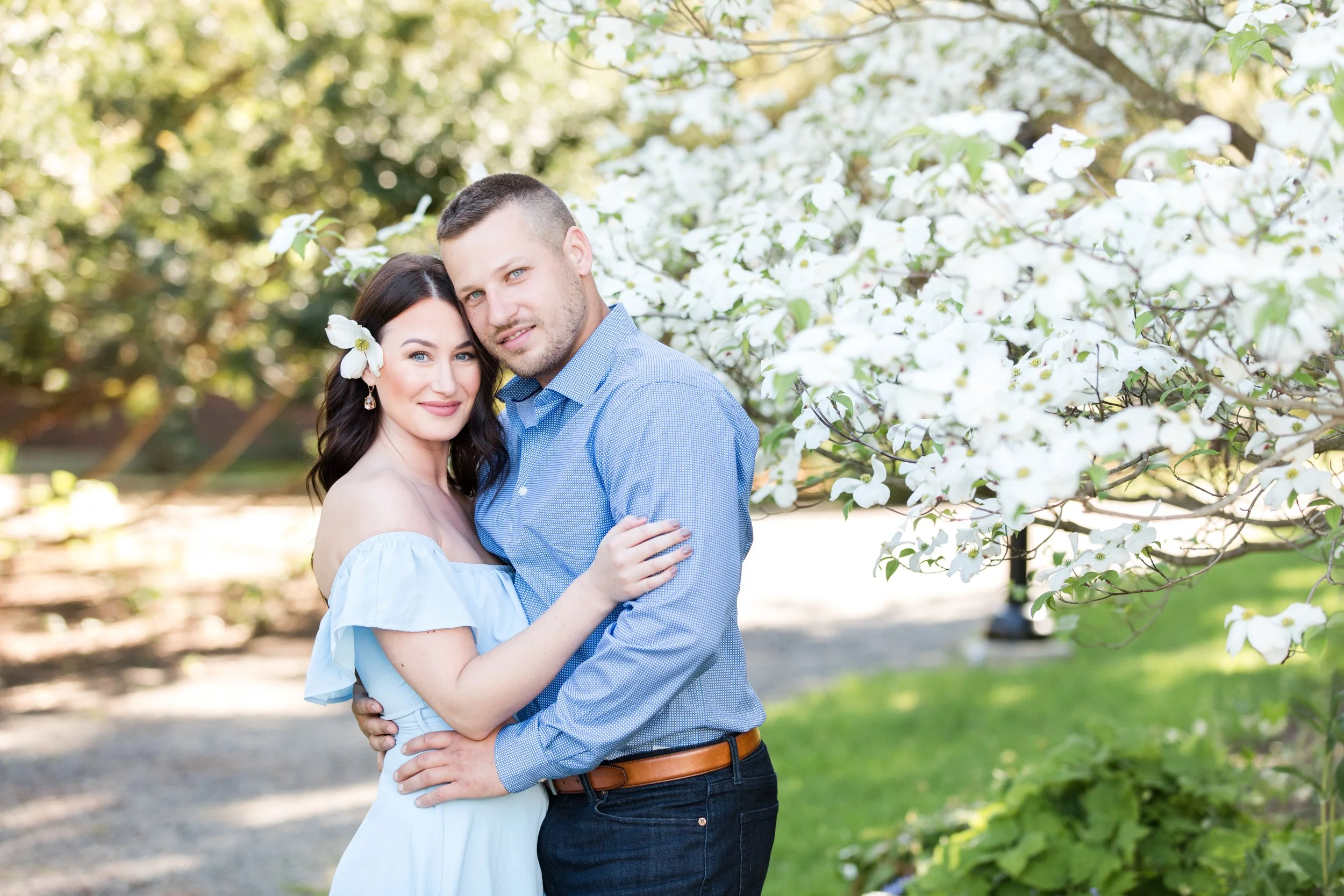 A young couple in a park during spring, embracing each other, with white blooming trees in the background, the woman has a flower in her hair and is wearing an off-shoulder light blue dress, the man is wearing a blue shirt and dark jeans.