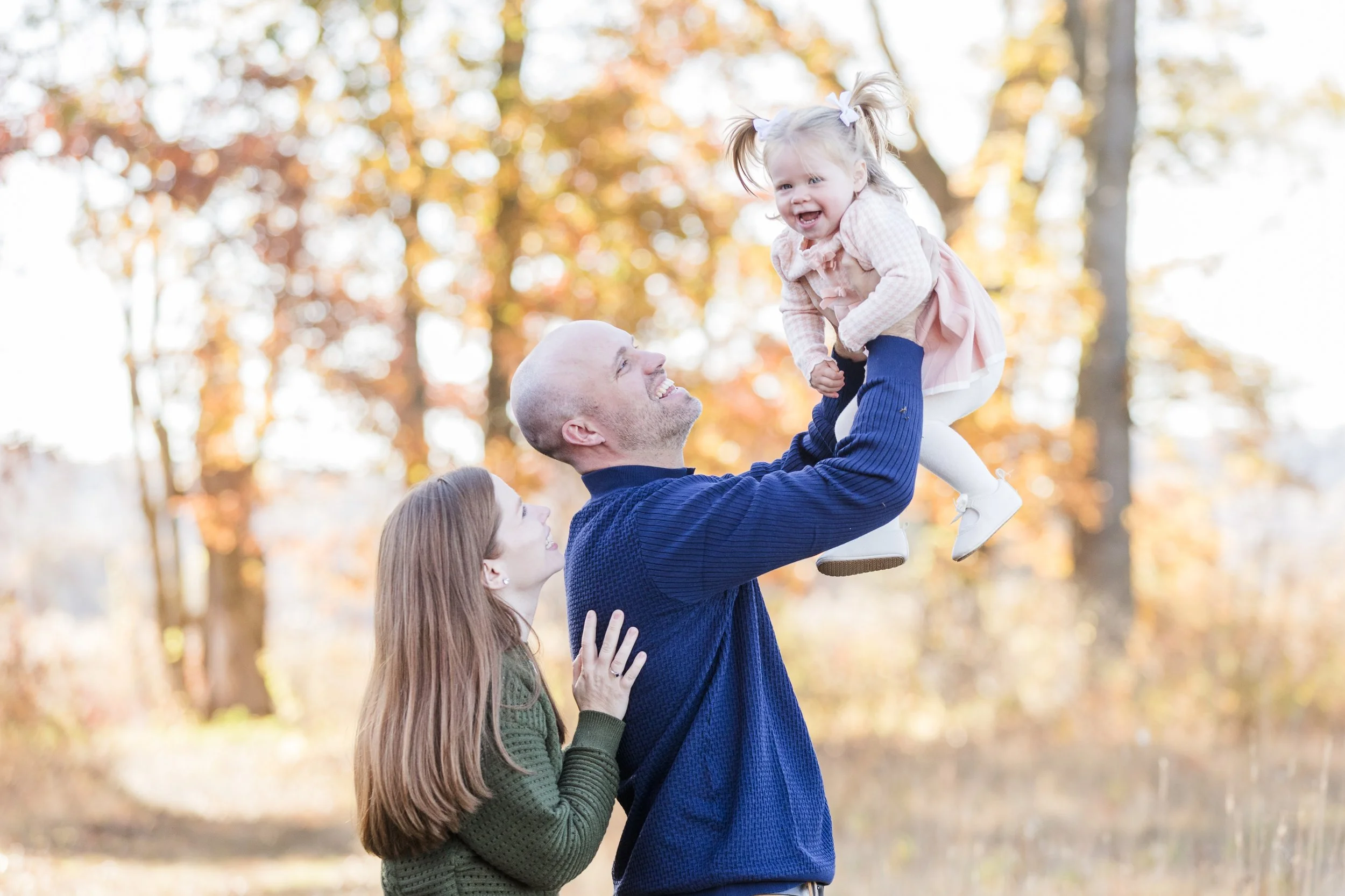 A man lifting a young girl into the air while a woman looks on affectionately in a park with orange and yellow autumn leaves.