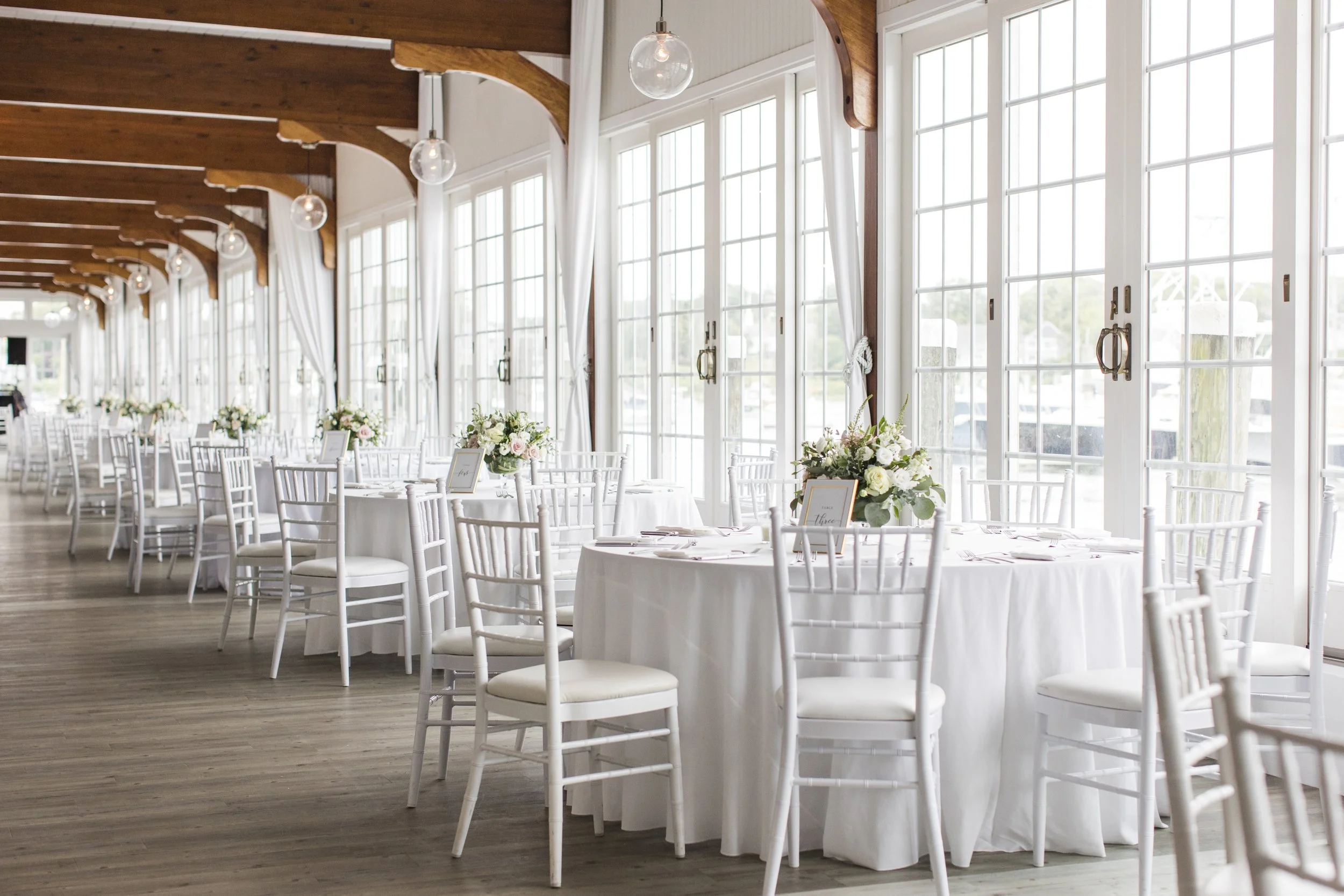 Elegant wedding reception area with white round tables, white chairs, floral centerpieces, and large windows with white curtains in a bright room with wooden beams.