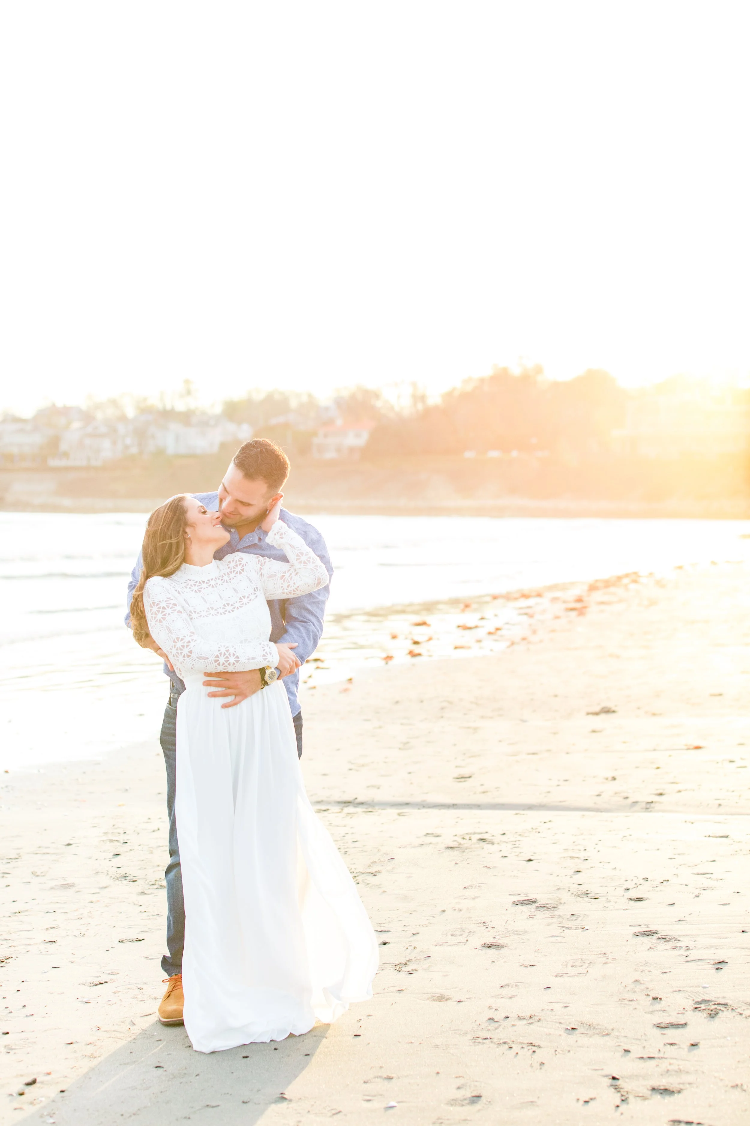 A couple embracing on a beach at sunset, the woman in a white dress and the man in a blue shirt, with the ocean and houses in the background.