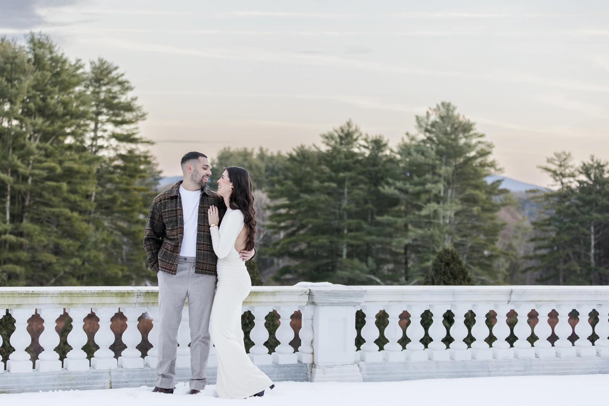 A couple standing on a snow-covered balcony with a forest of pine trees in the background during winter.