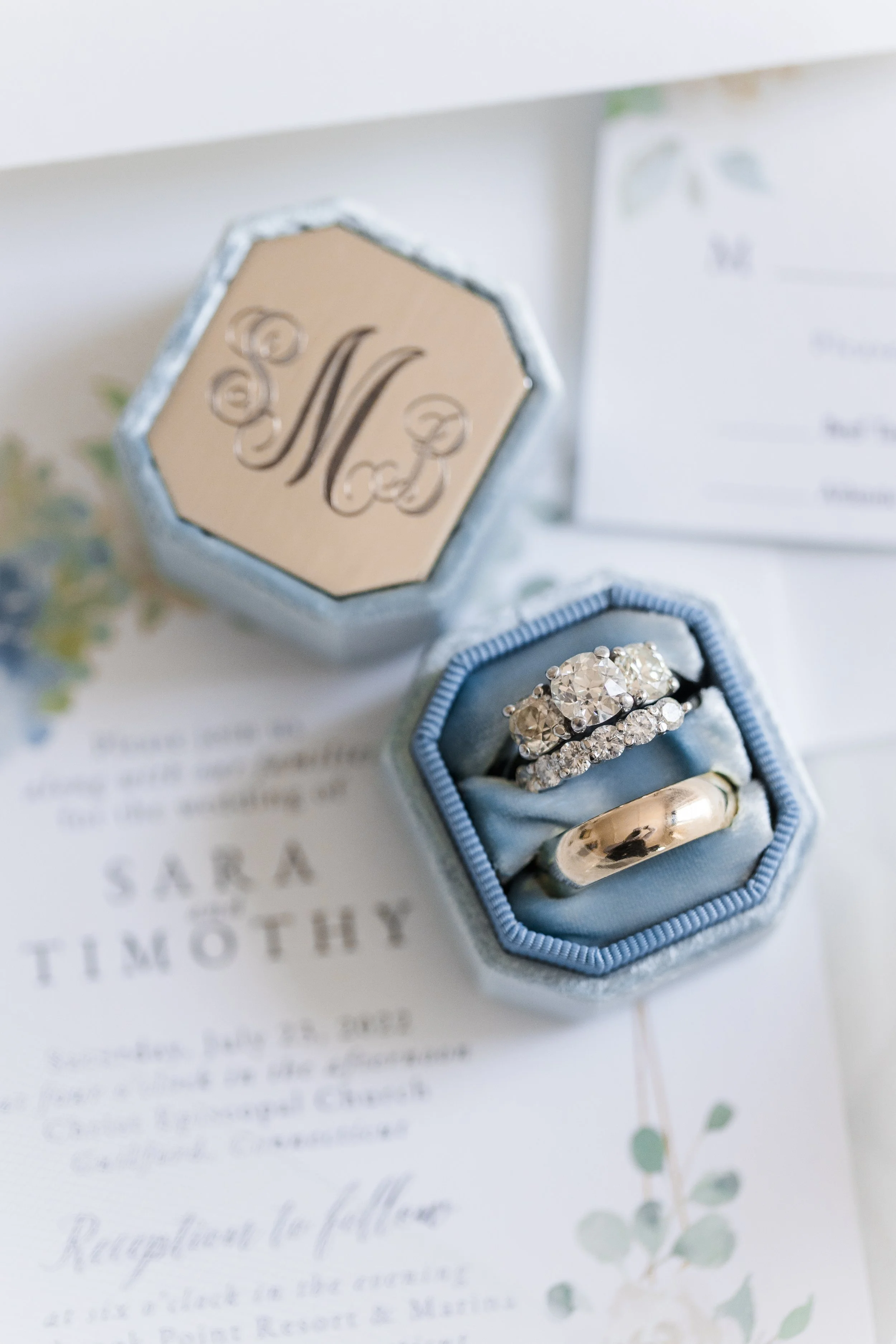 Close-up of a jewelry box with wedding rings and diamond rings inside, placed on a wedding invitation.