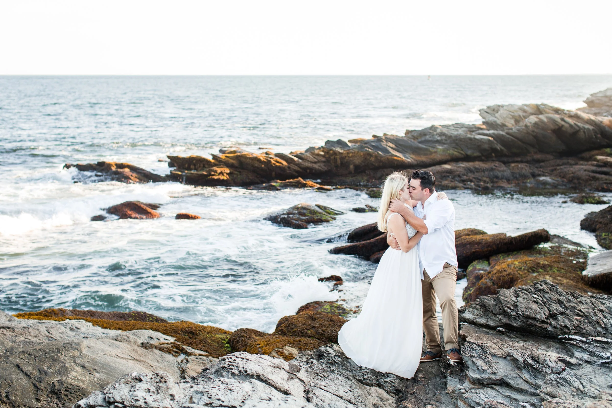 A couple kissing on rocks by the ocean, with waves crashing around them and a clear sky in the background.