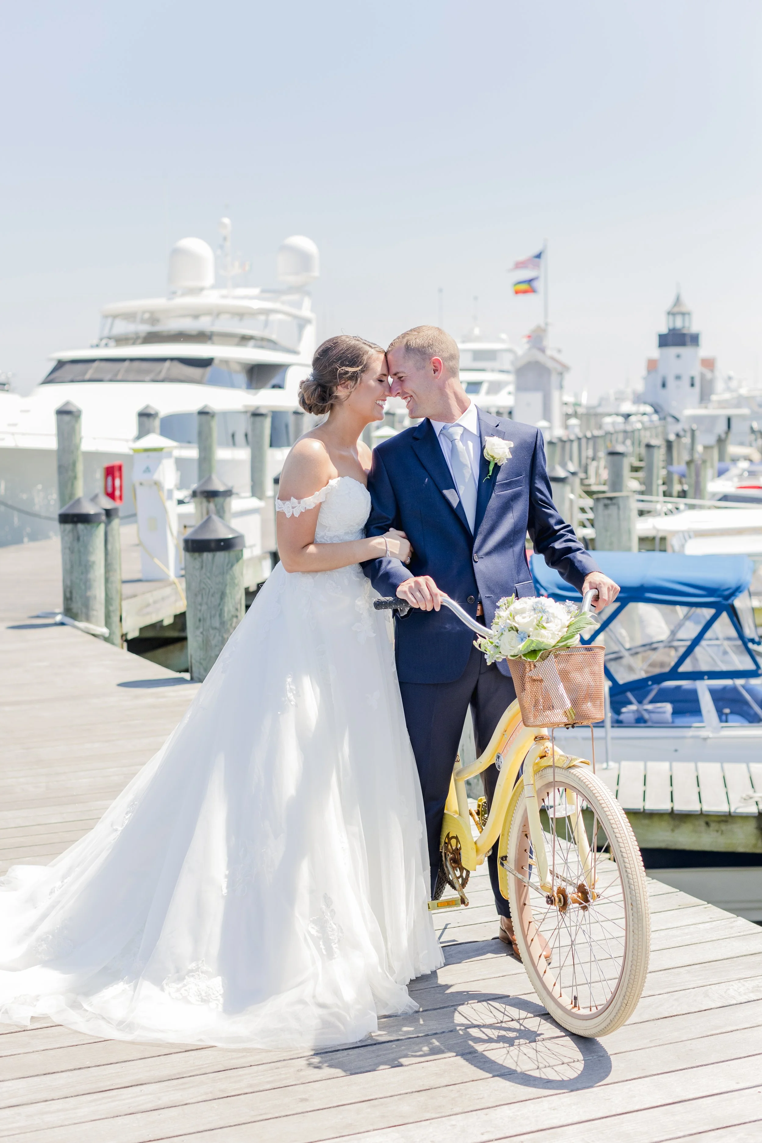 A bride and groom in wedding attire standing on a pier near boats, with the groom holding a bicycle with a flower basket, under a clear sky.