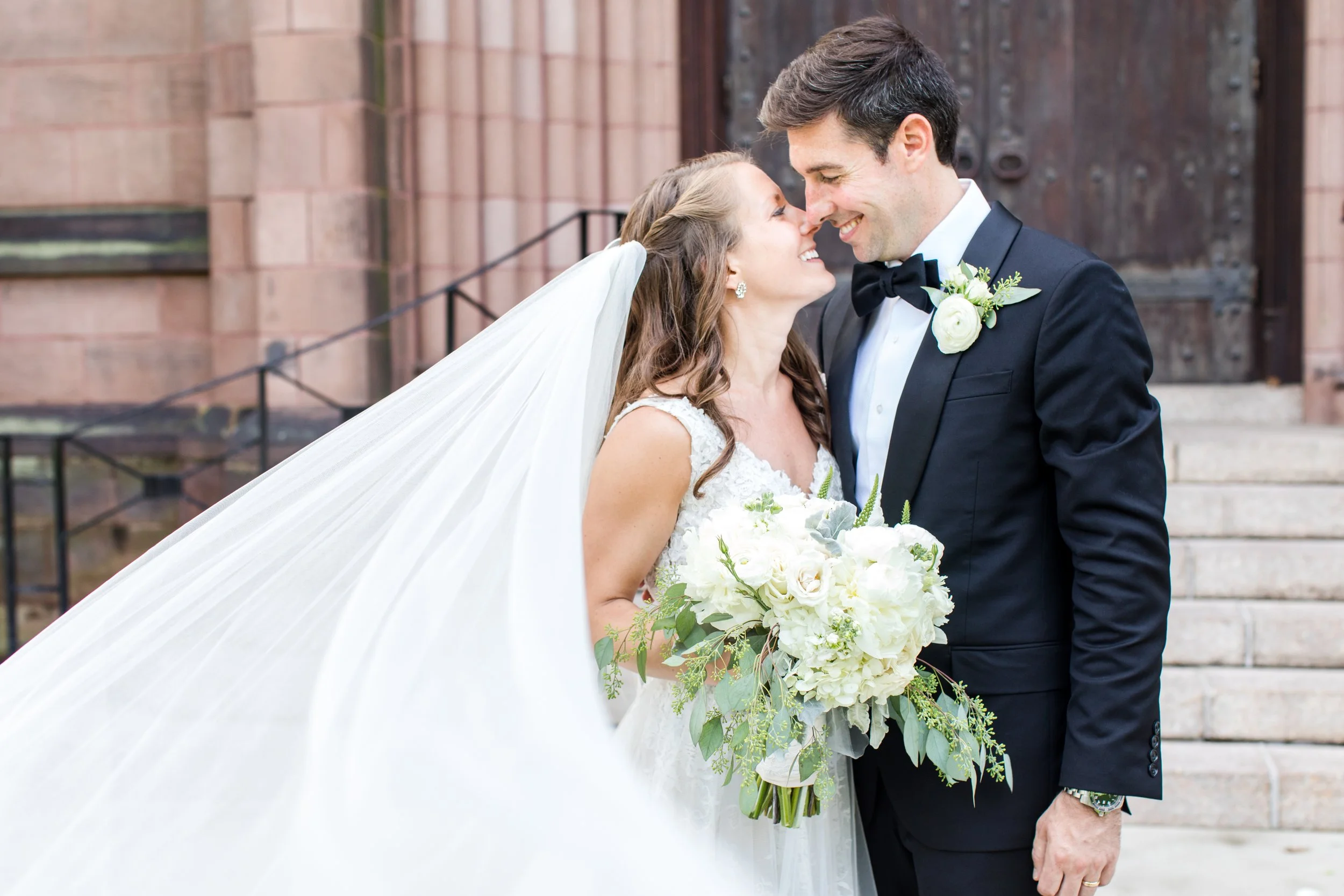 Bride and groom smiling closely on wedding day, bride holding a bouquet of white flowers, bride wearing a white wedding dress with veil, groom in a black tuxedo with bow tie and white boutonniere, standing outside near stone steps.