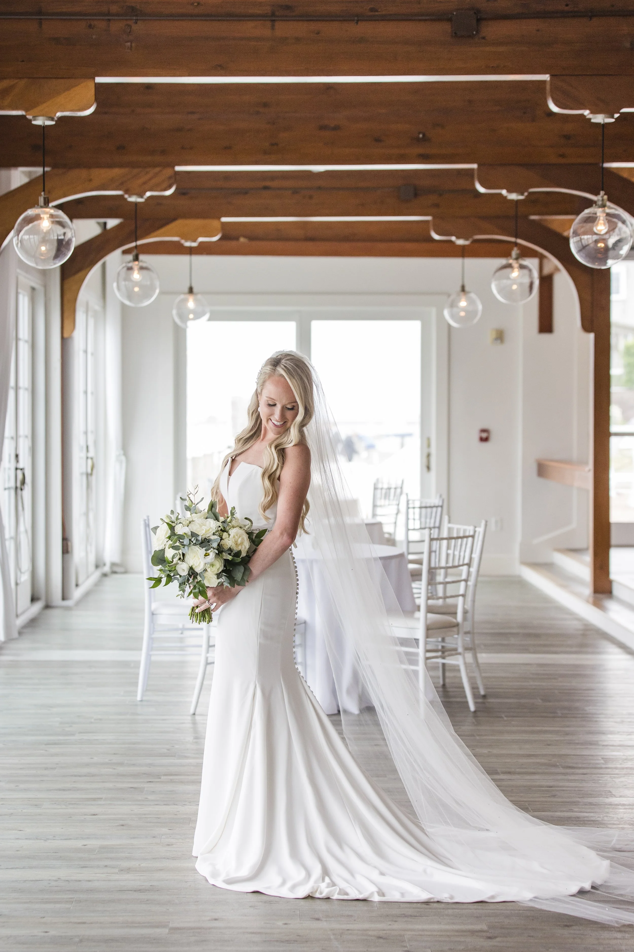 A bride with long blonde hair in a white wedding gown holding a bouquet of white and green flowers, standing inside a bright, modern wedding venue with wooden beams and hanging light fixtures.