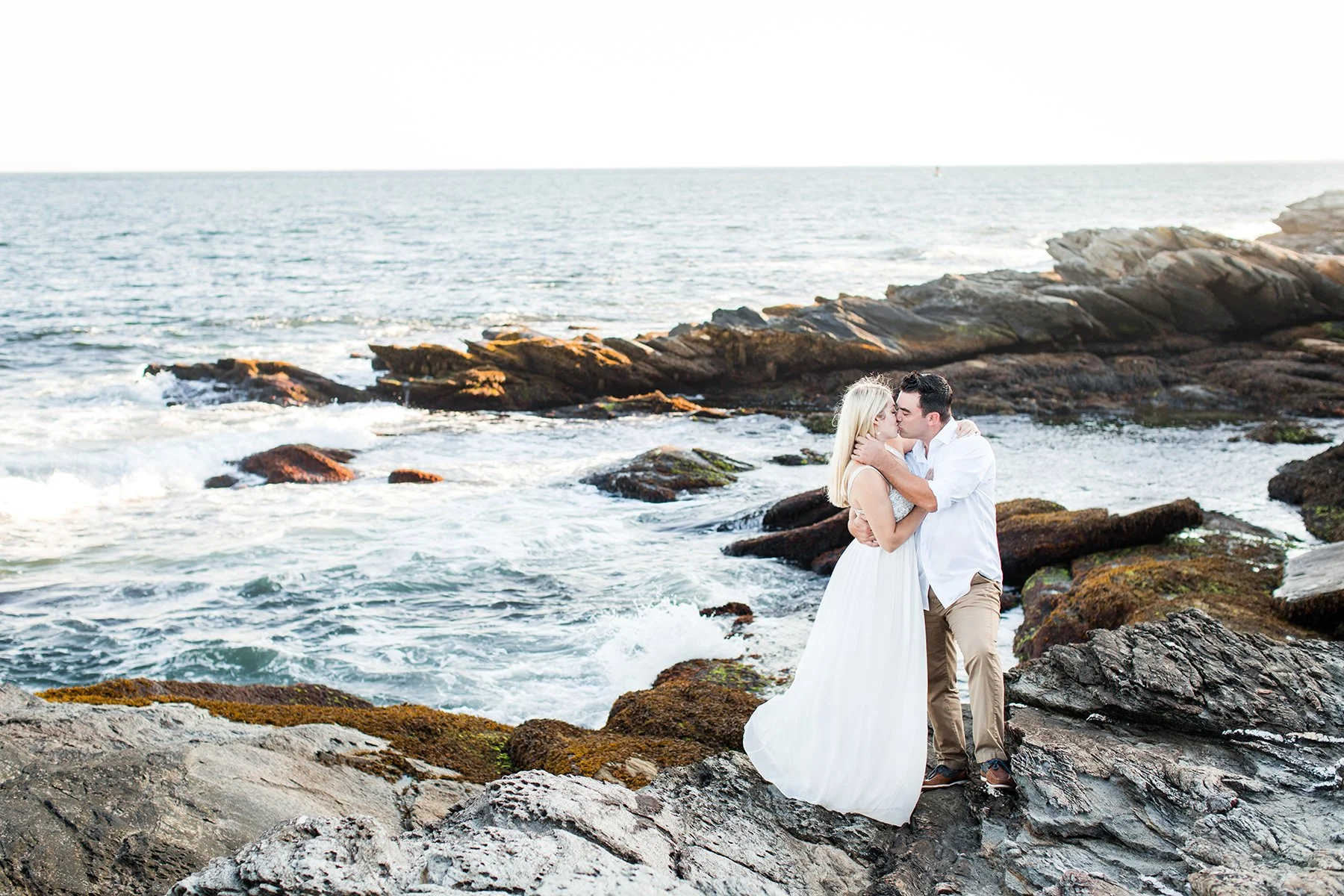 Couple sharing a kiss on rocky shoreline near the ocean during sunset.