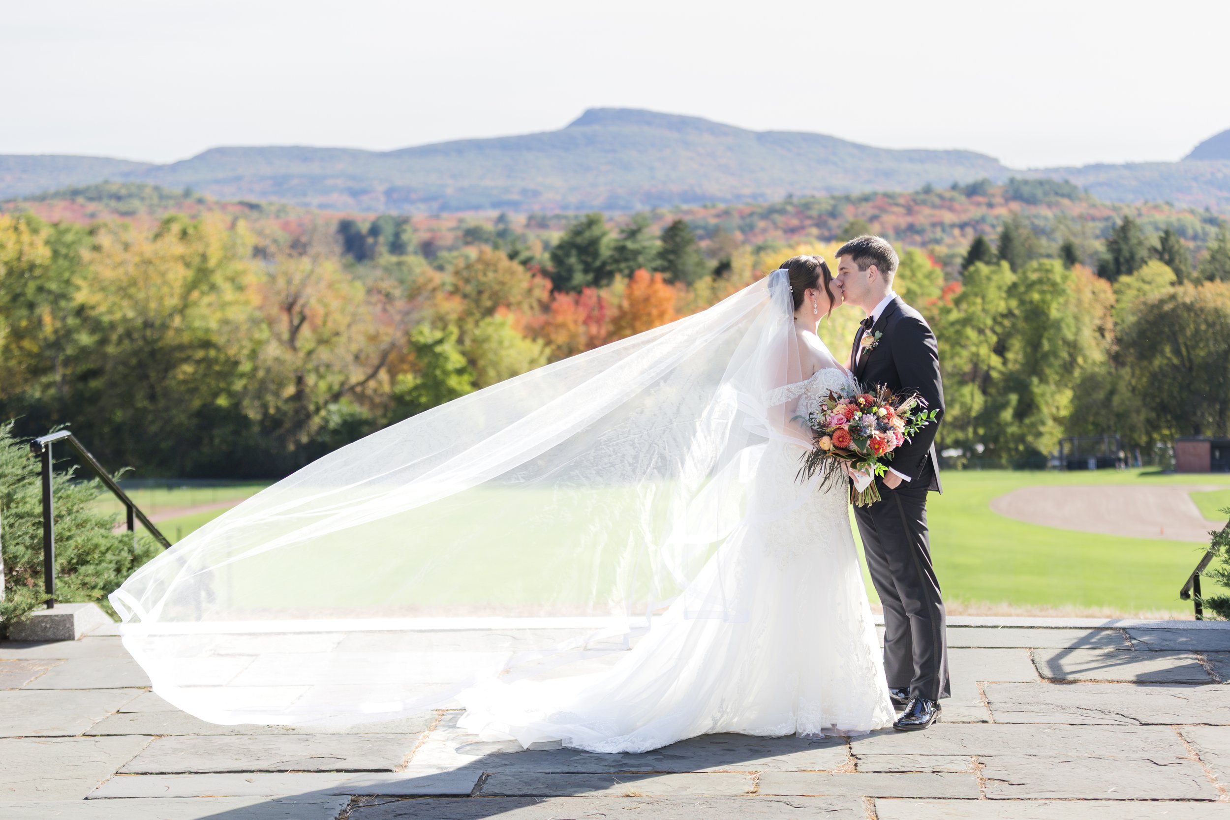 A bride and groom share a kiss outdoors on a stone pathway, with the bride holding a bouquet of flowers. She wears a white wedding dress with a long veil, and the groom wears a black tuxedo. Trees with fall foliage and mountains are in the background