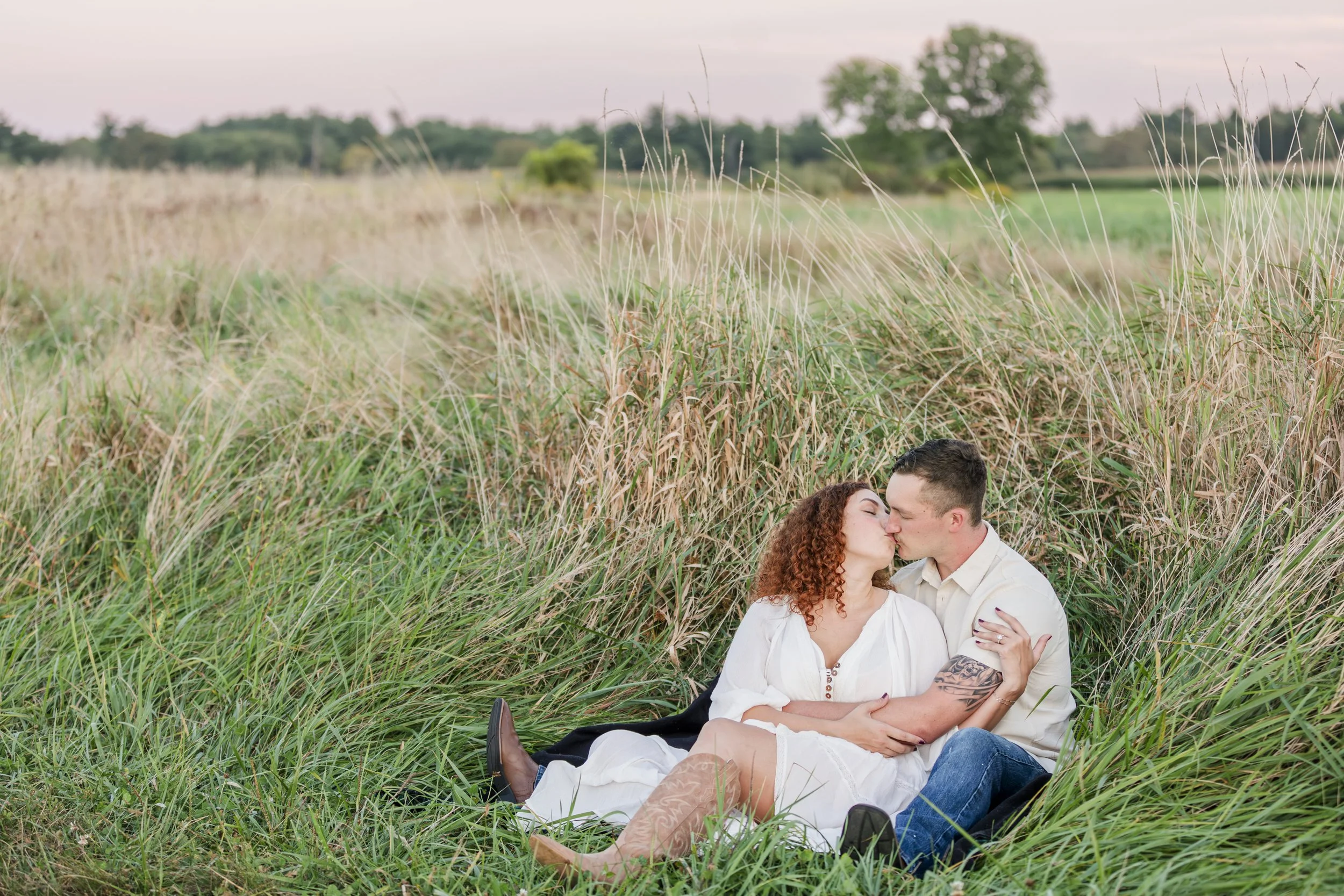A couple sitting and kissing in a field of tall grass during sunset.