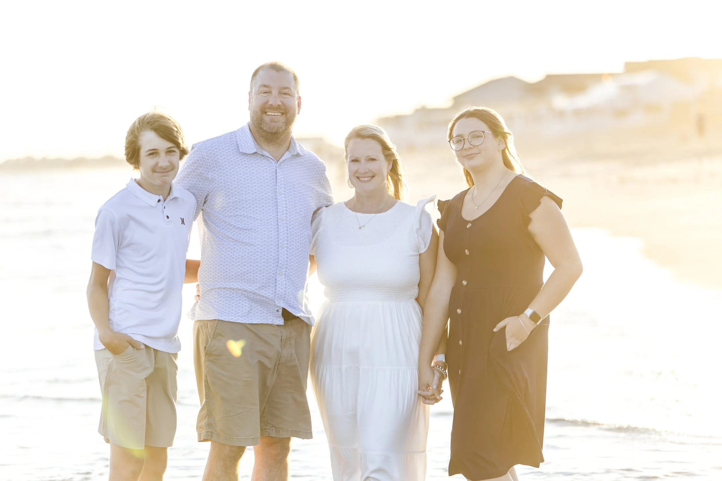 Family of four standing on the beach at sunset, smiling and holding hands.