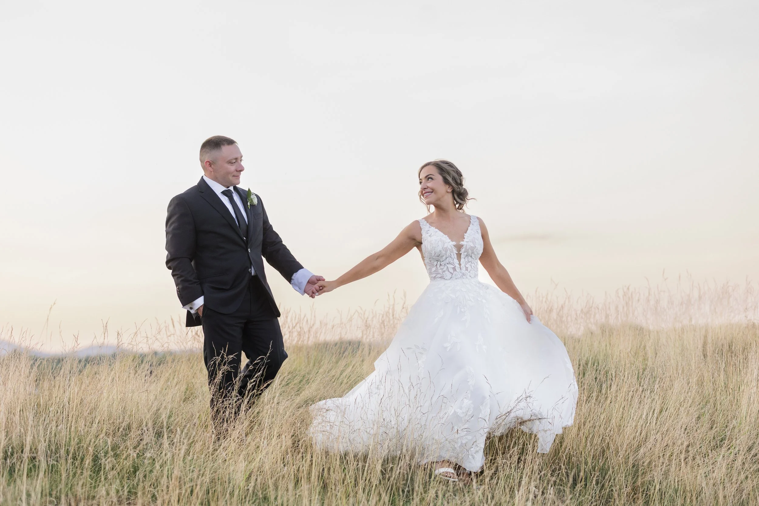A bride and groom walking hand in hand through a grassy field during sunset, smiling at each other.
