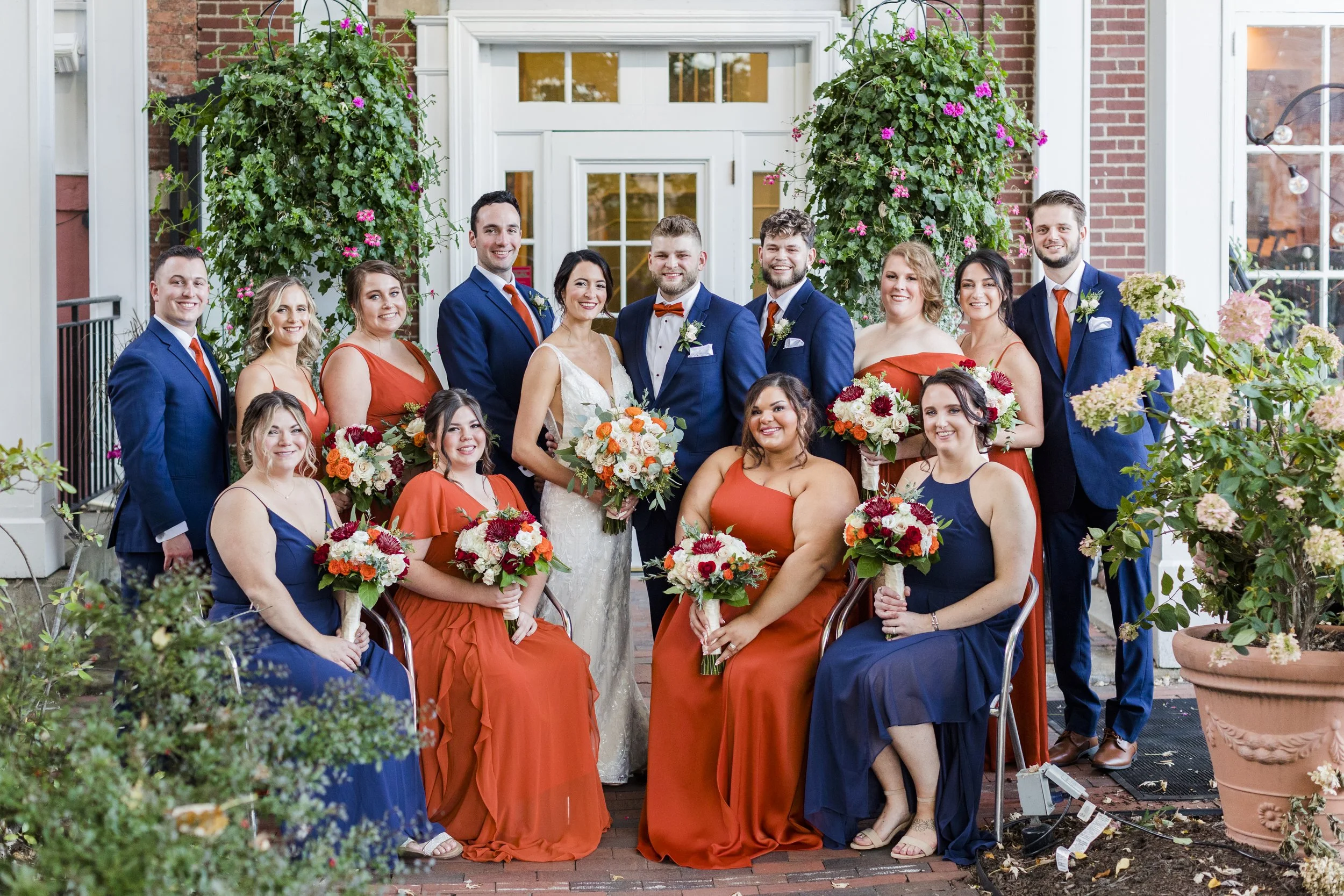 Group of wedding party members, including the bride in a white dress, groom in a blue suit, bridesmaids in orange and navy dresses, and groomsmen in blue suits, posing outdoors in front of a brick building with flowering plants and greenery.