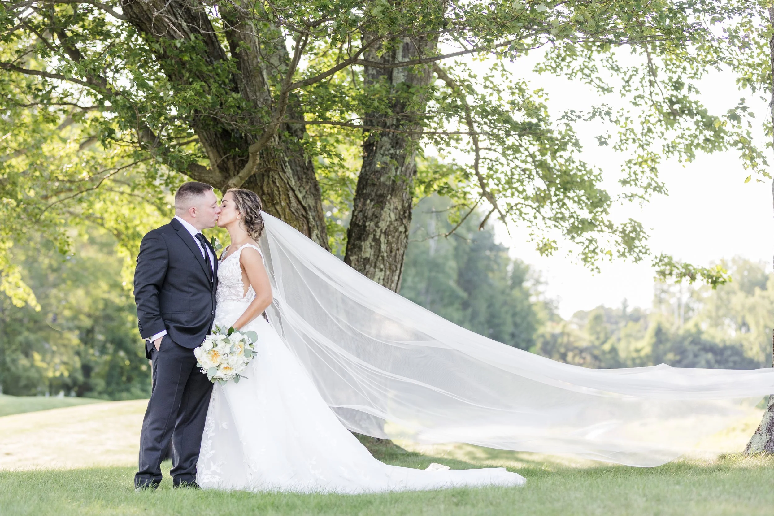 A bride and groom kiss outdoors under a large tree, the bride wearing a white wedding dress with a long veil and holding a bouquet, and the groom in a dark suit and tie, in a lush green park setting.