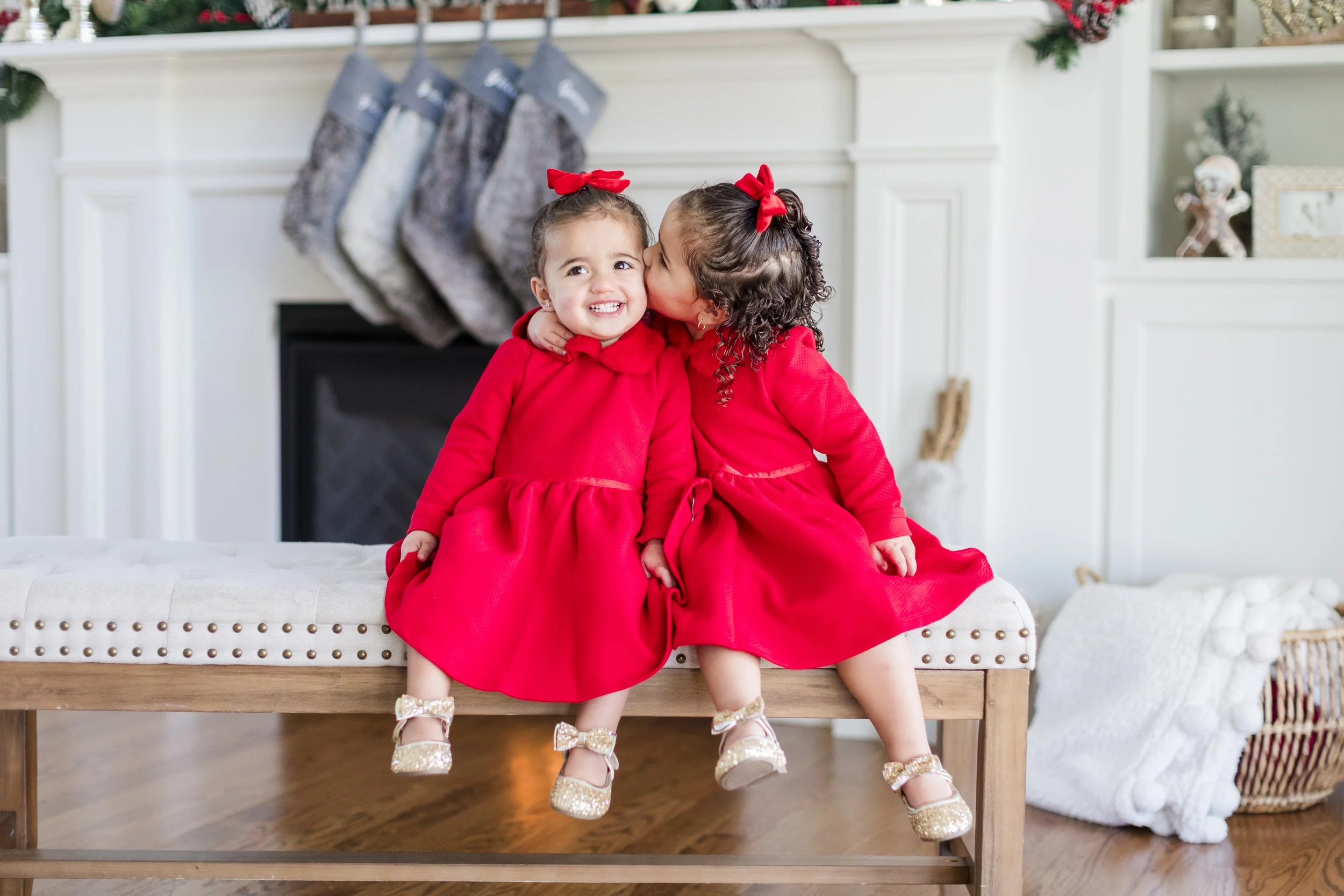 Two young girls in matching red dresses and bows sitting on a bench, one is kissing the other on the cheek inside a decorated Christmas living room.