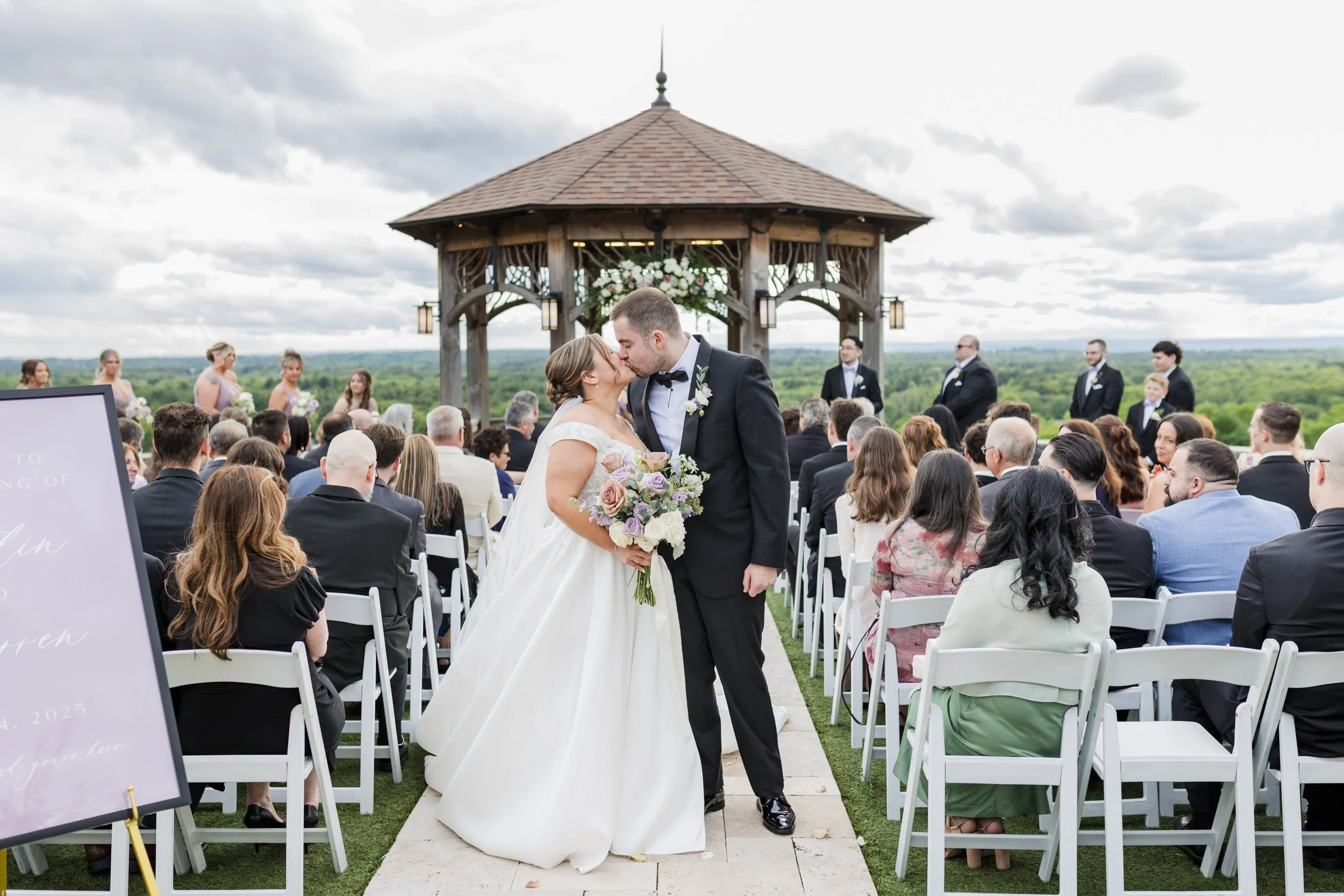 A bride and groom kissing at an outdoor wedding ceremony on a cloudy day, with guests seated on either side and the officiant and wedding party in the background, under a wooden gazebo with a scenic green landscape.