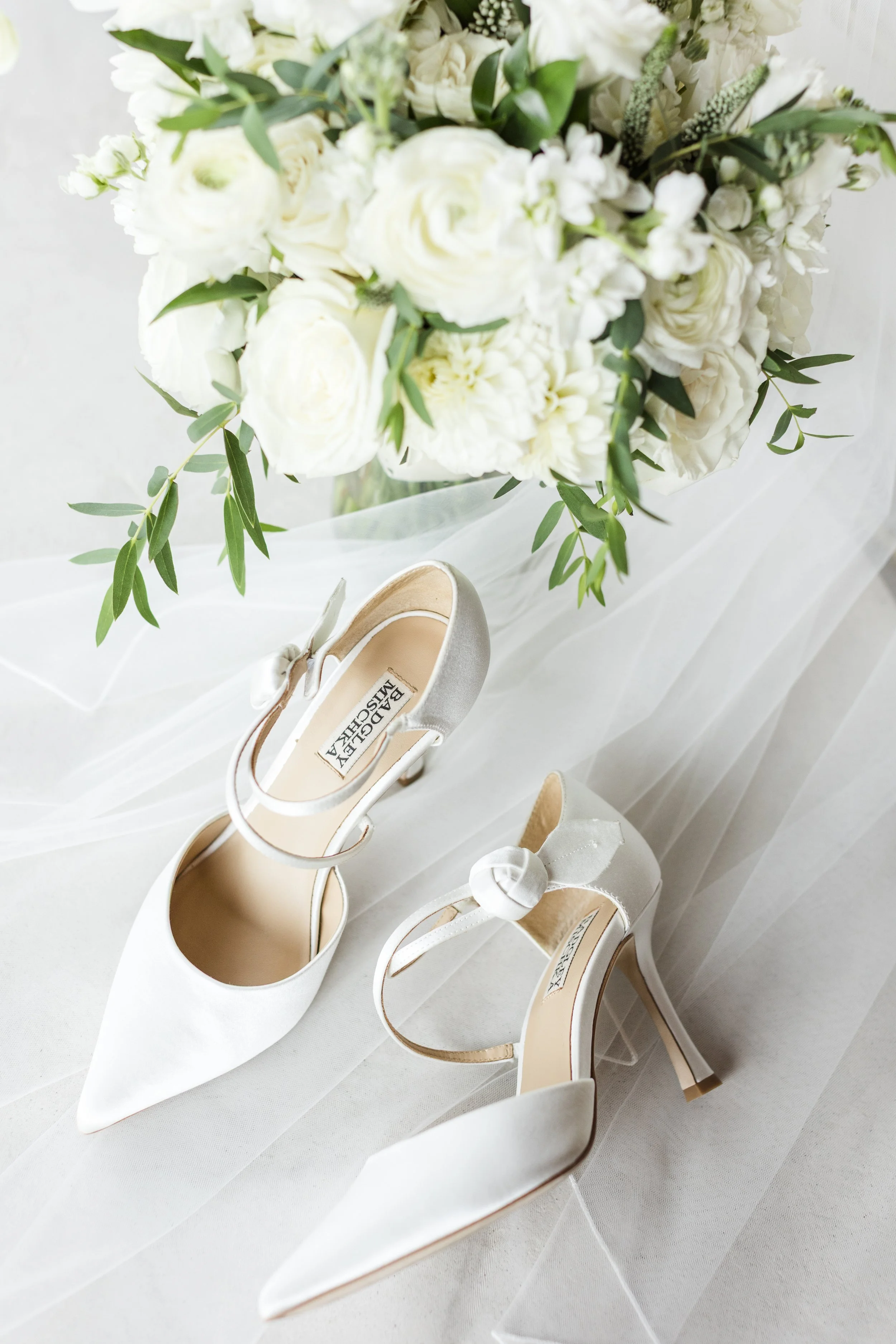 A pair of white high-heeled bridal shoes with satin ribbons and a bouquet of white roses, hydrangeas, and greenery.