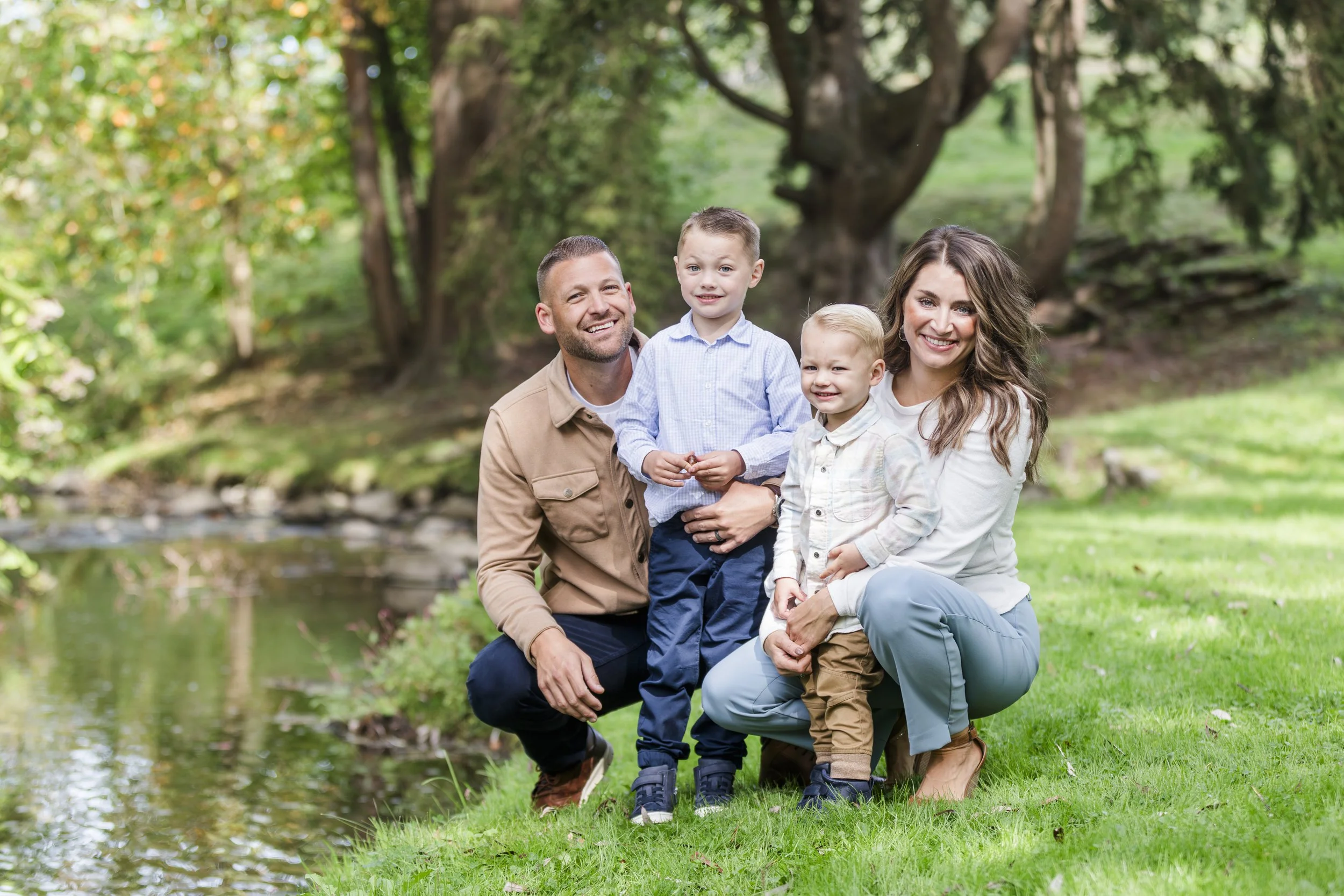 A family of four posing together outdoors near a pond, with trees in the background. The parents are kneeling, smiling, and the two young boys are standing between them, also smiling.