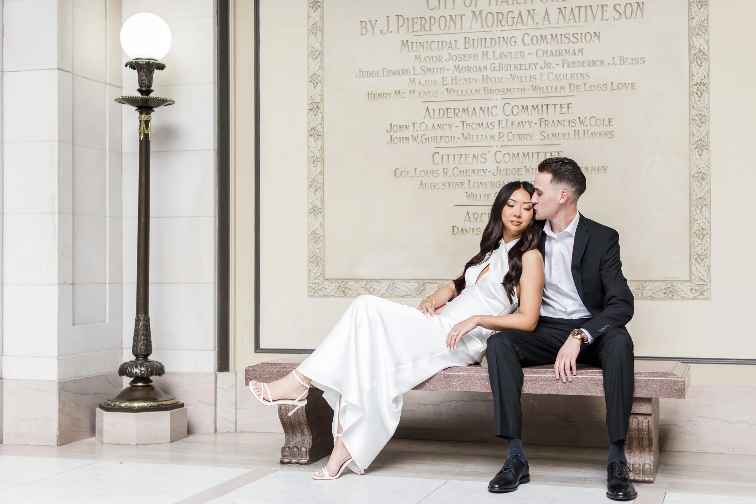A couple dressed in formal attire sitting on a bench inside a building with carved wall details and historical plaques, the woman in a white dress and the man in a black suit, sharing a tender moment.
