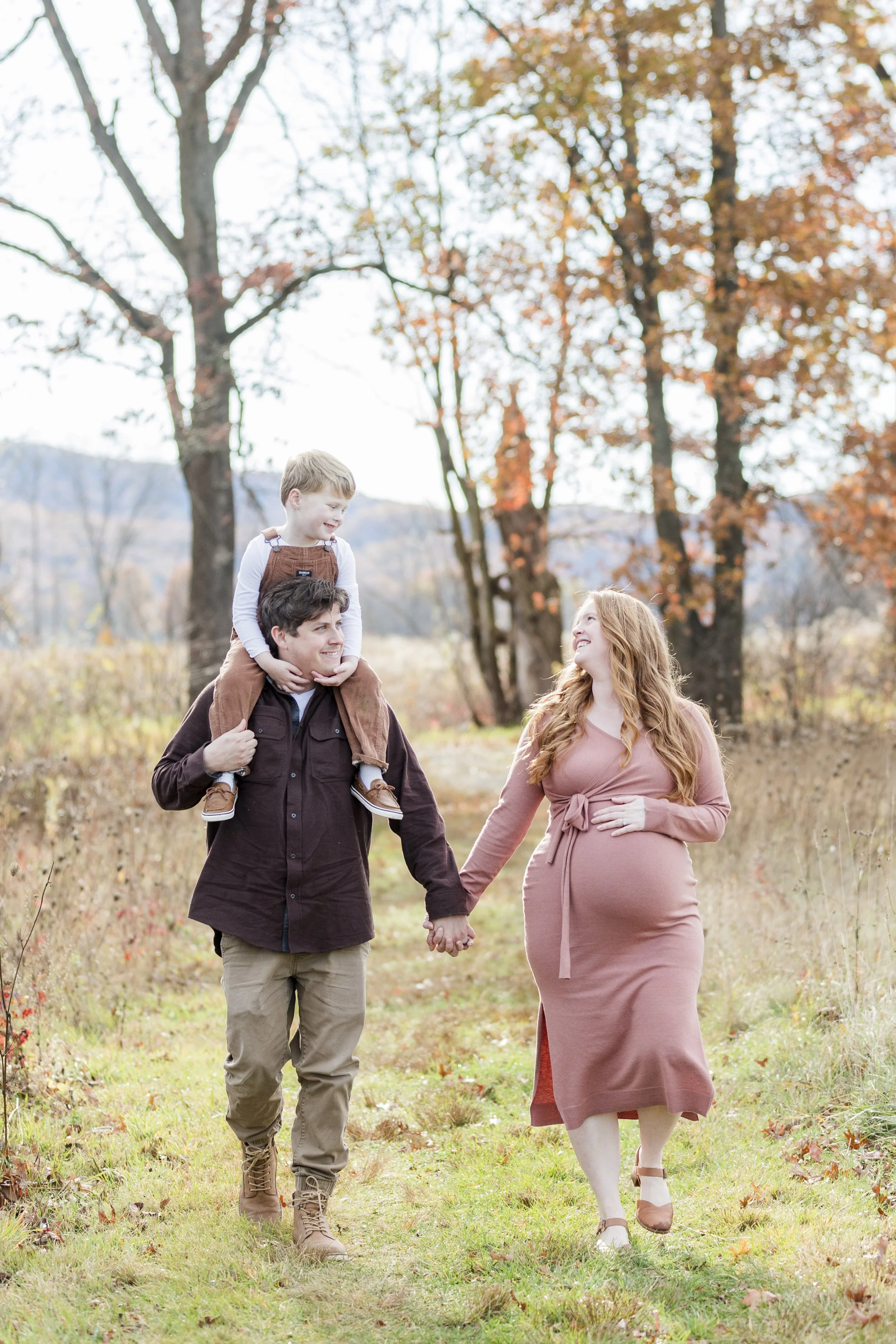 A family of three walking outdoors in an autumn landscape: a man carrying a young boy on his shoulders, and a woman holding the man's hand. The woman appears pregnant and is smiling.
