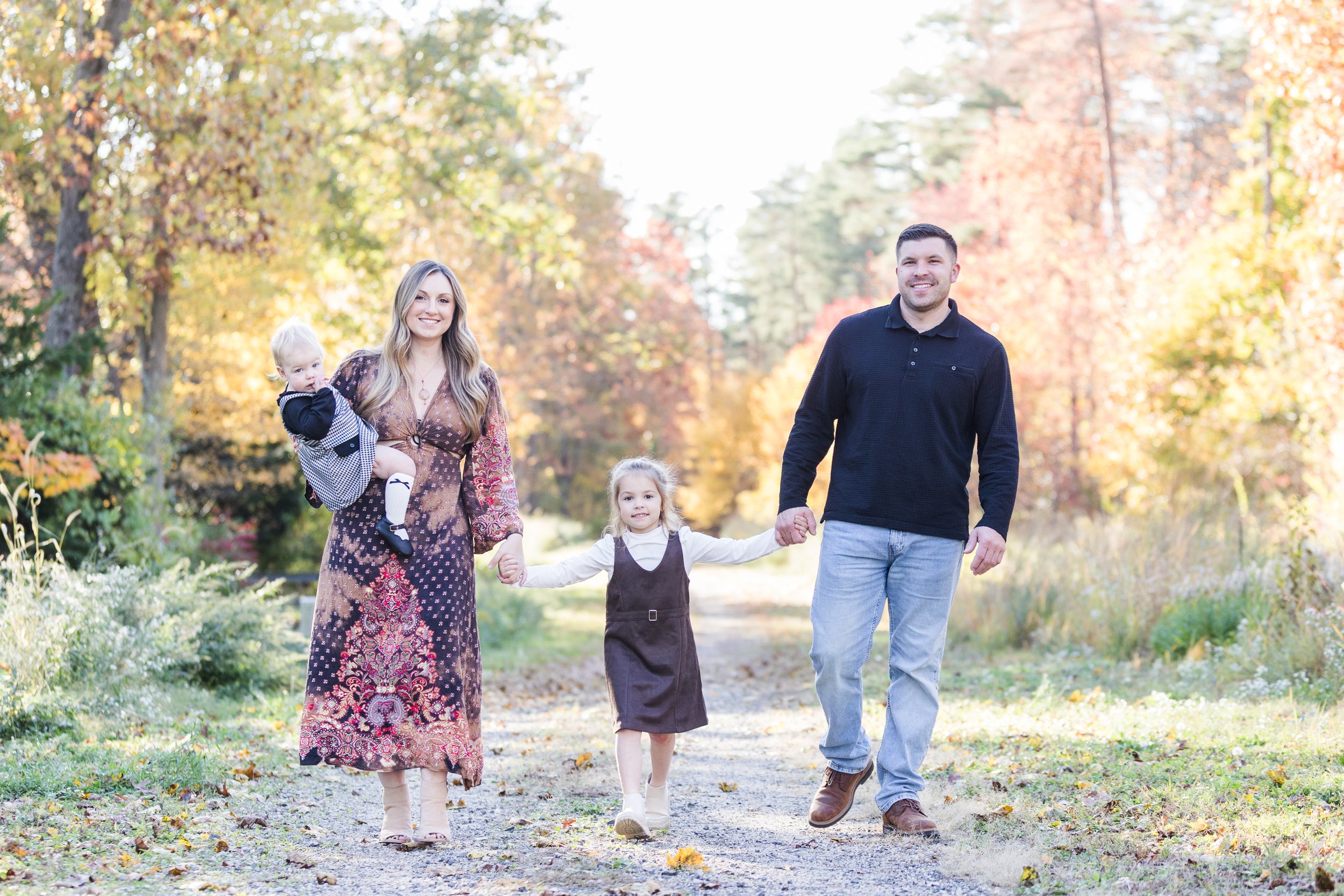 A smiling family of four walking hand-in-hand outdoors on a fall day, surrounded by colorful autumn trees.