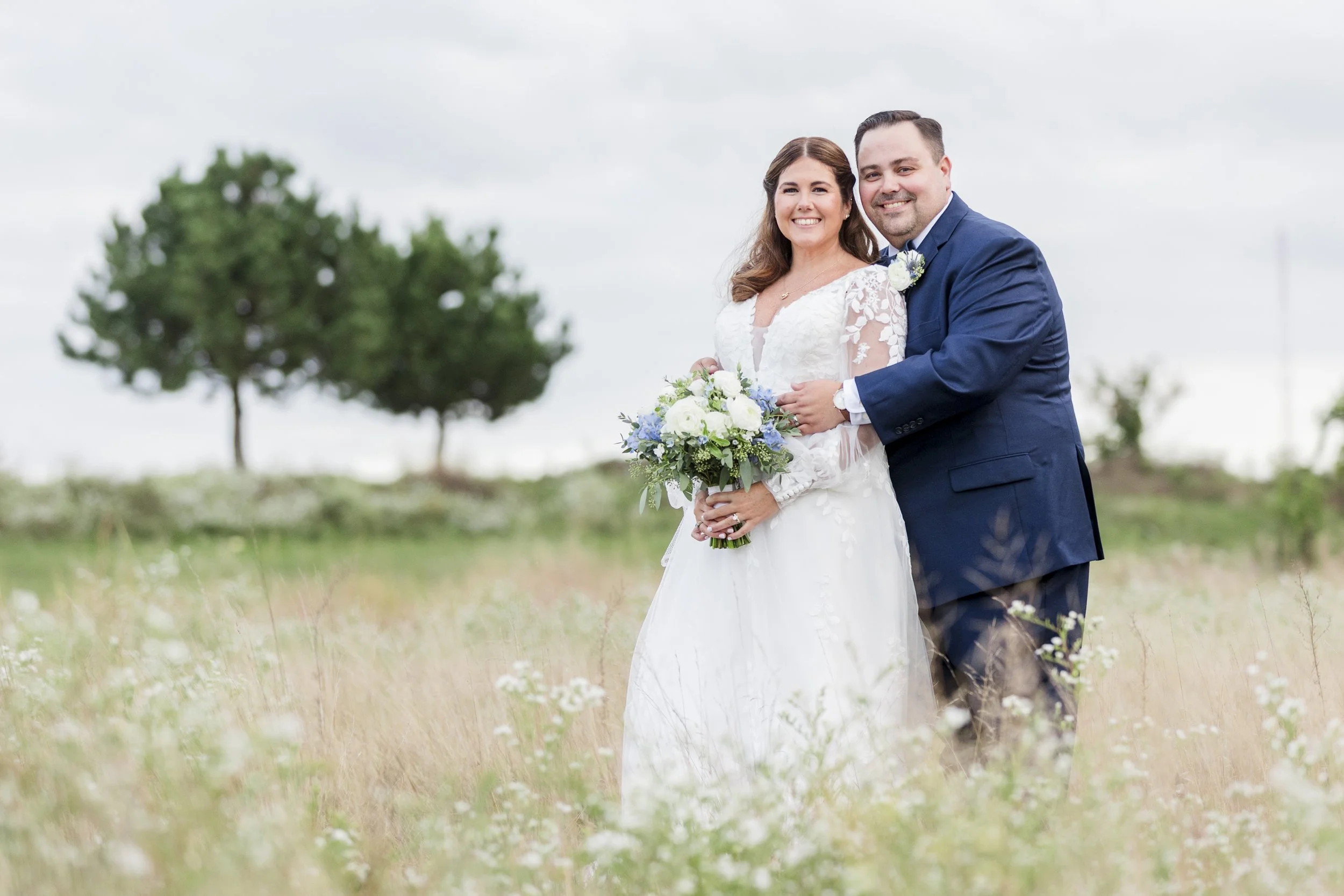 A newlywed couple standing in a field of tall grass and wildflowers on a cloudy day. The bride is holding a bouquet and wearing a white lace wedding dress, and the groom is in a navy blue suit. They are smiling and posing together.