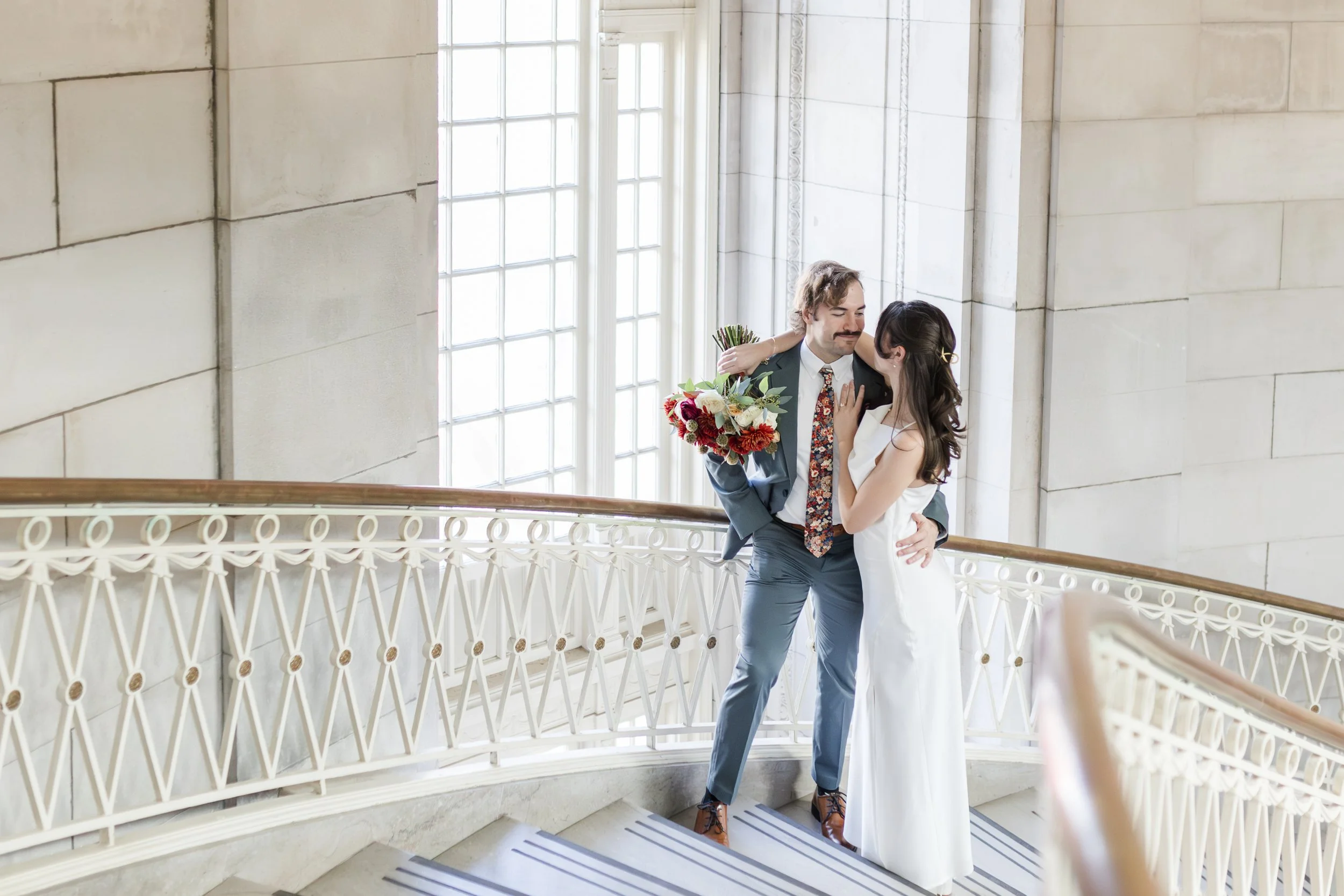 A couple dressed in wedding attire standing on a staircase, embracing and smiling at each other. The man is holding a bouquet of flowers, and they are in a bright, elegant indoor setting with large windows.