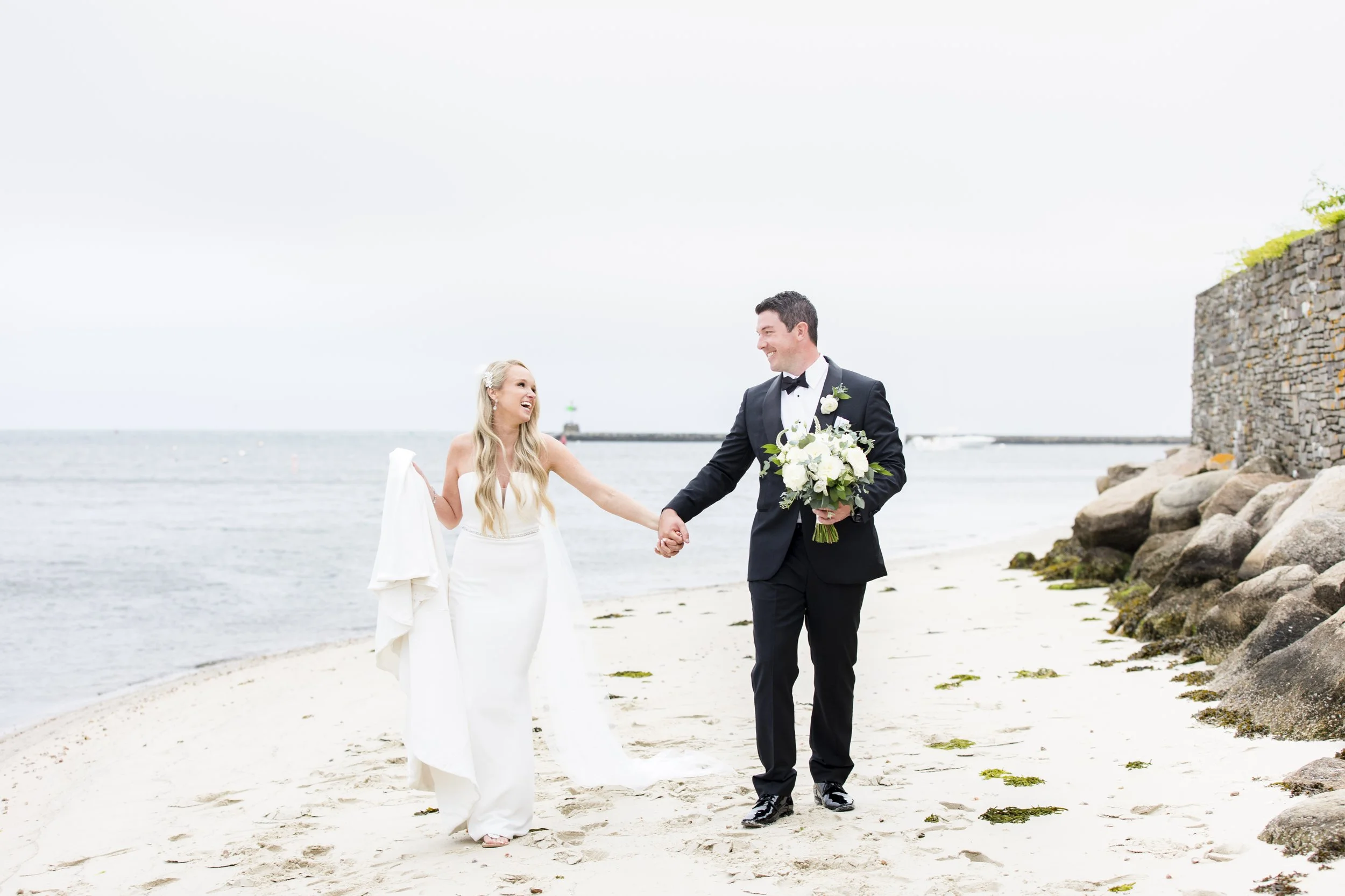 A bride and groom walking hand in hand on a beach, smiling at each other. The bride is dressed in a white wedding gown holding a white cloth, and the groom in a black tuxedo holding a bouquet of white flowers. The background includes the ocean, rocks