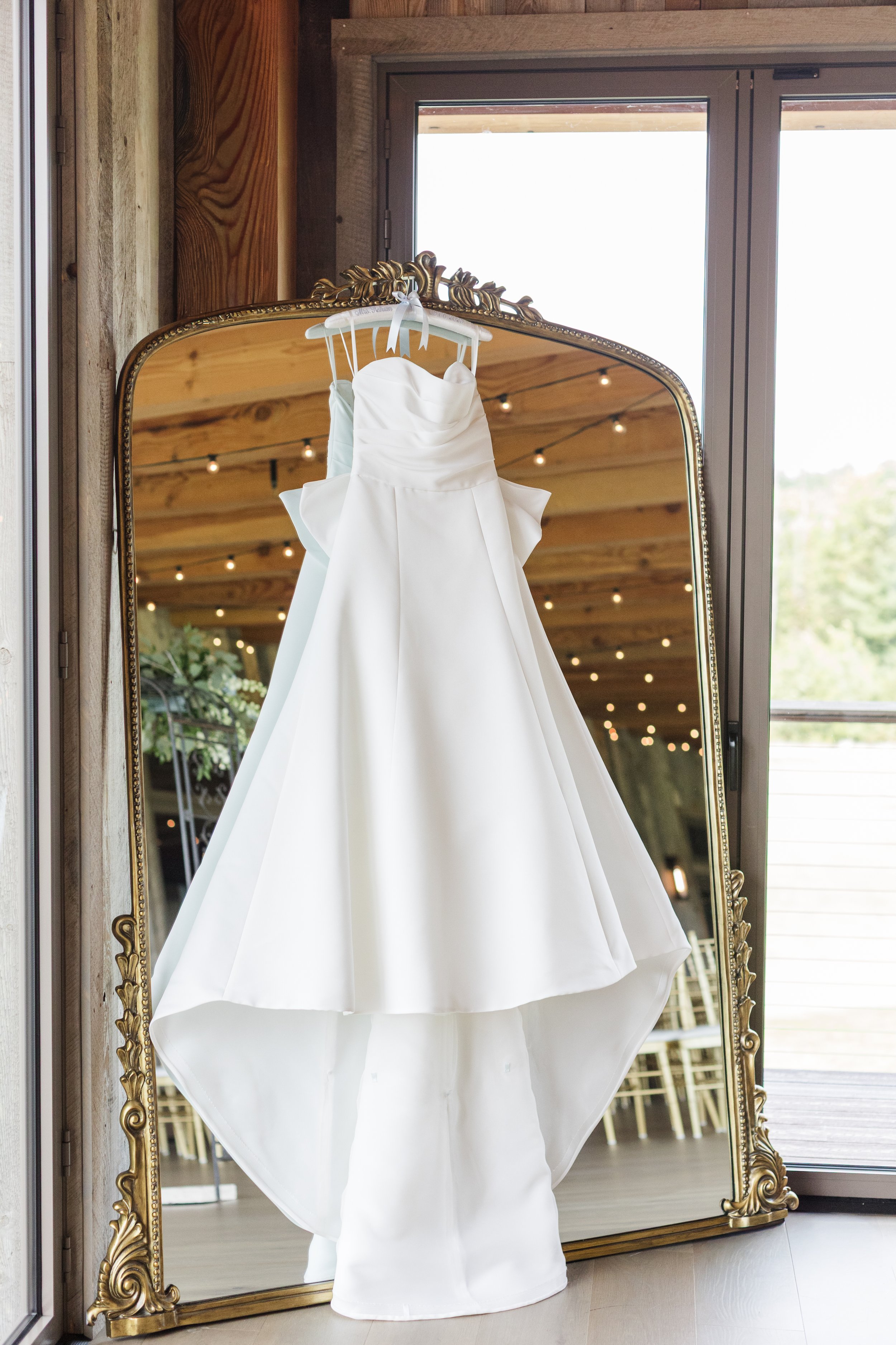 A white wedding dress hanging on a white hanger in front of a large ornate mirror, inside a wooden room with a window showing an outdoor deck and greenery.