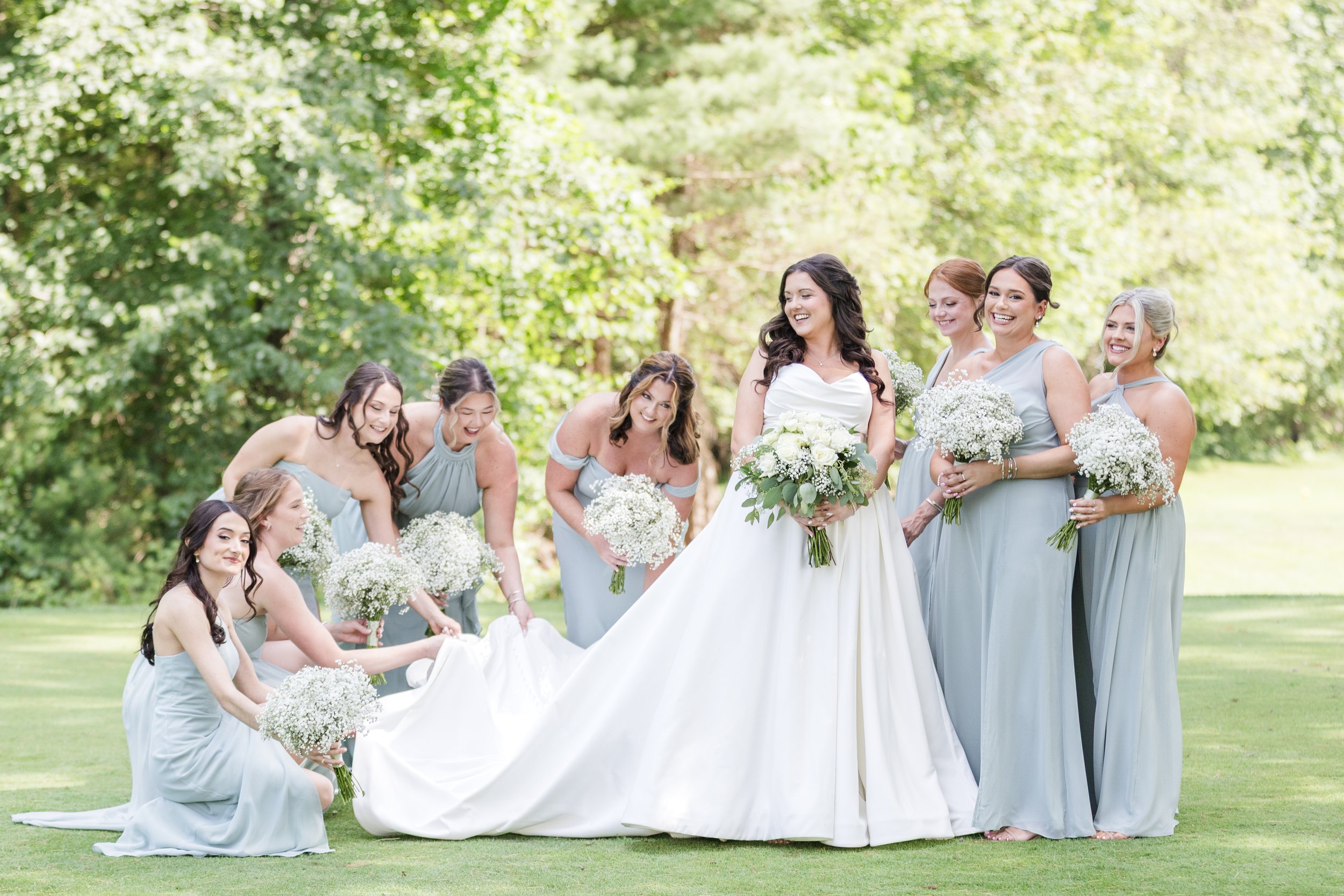 A bride in a white wedding dress surrounded by her bridesmaids in light blue dresses, all holding white bouquets, on a lush green lawn with trees in the background.
