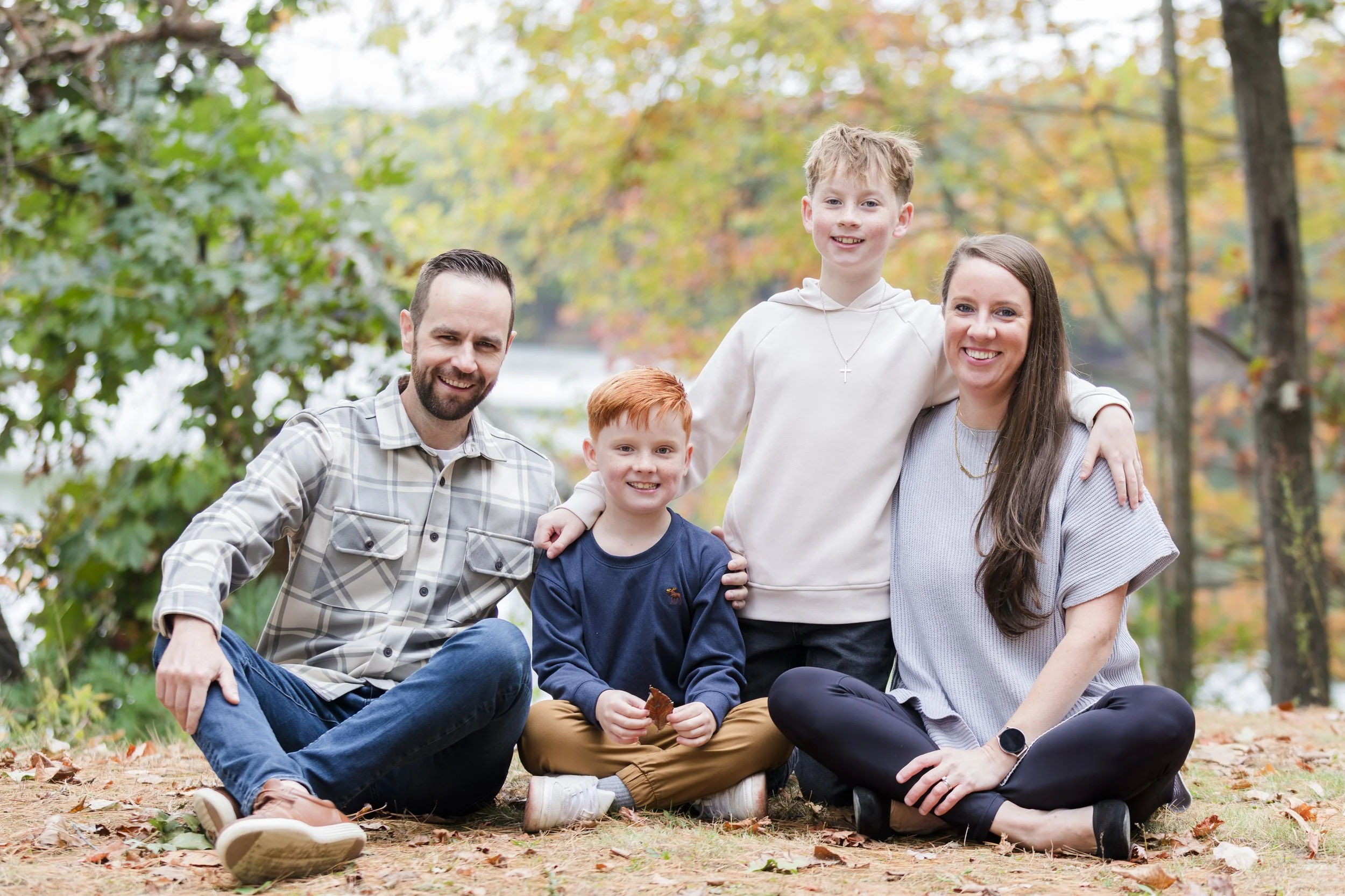 Family of four sitting and smiling on the ground in a park with fallen leaves, surrounded by trees with autumn foliage.