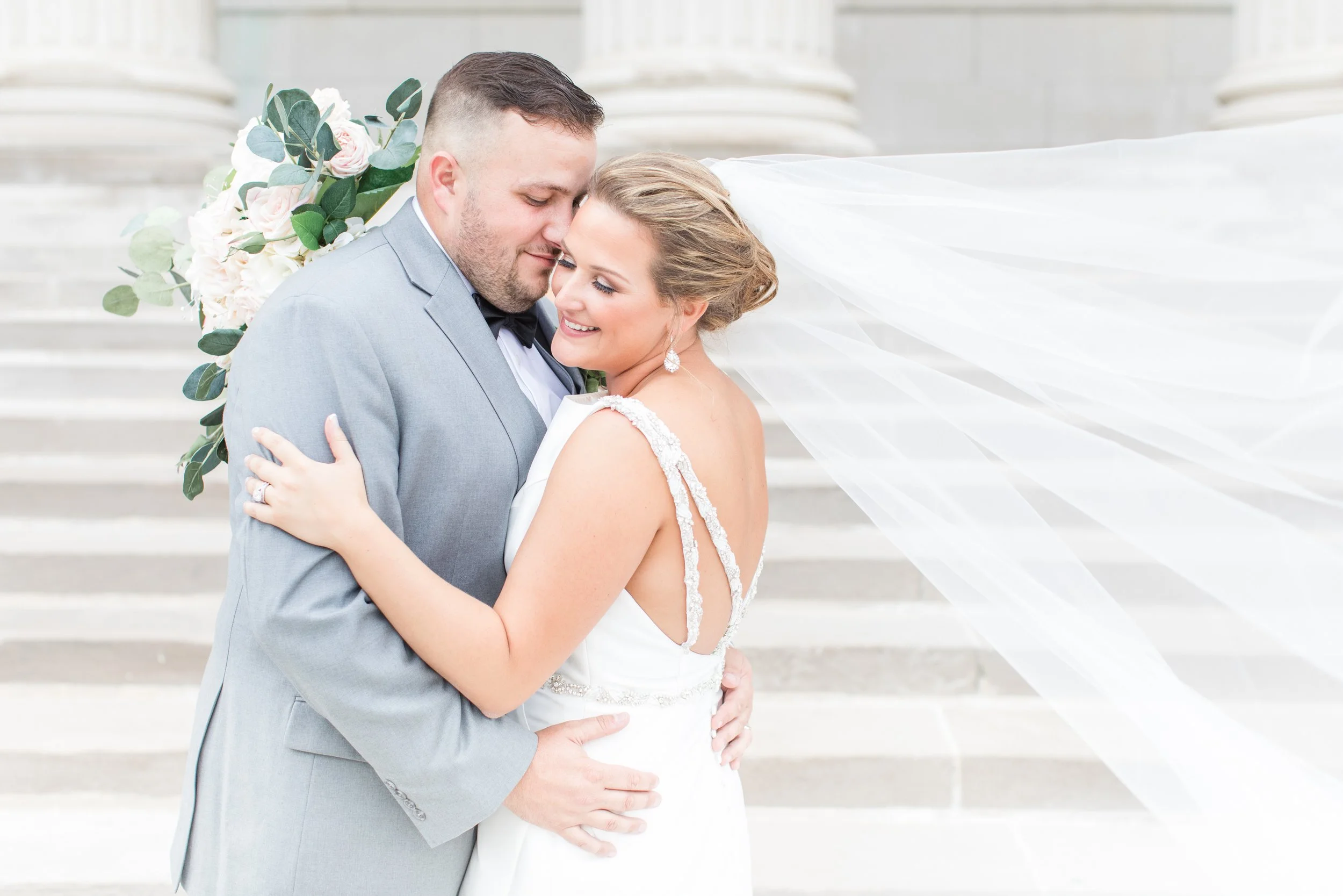 A bride and groom embracing in front of a staircase, with the bride's veil flowing in the wind and holding a bouquet of flowers behind them.
