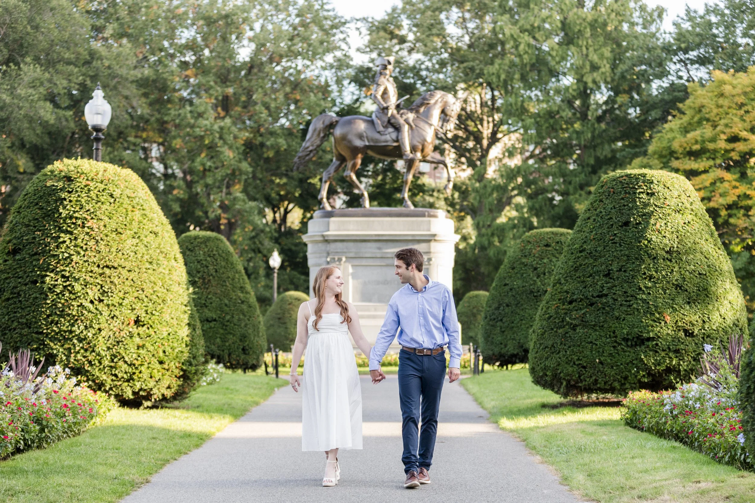 A young couple holding hands and walking in a park with green bushes, trees, flowers, and a bronze statue of a person on a horse in the background.