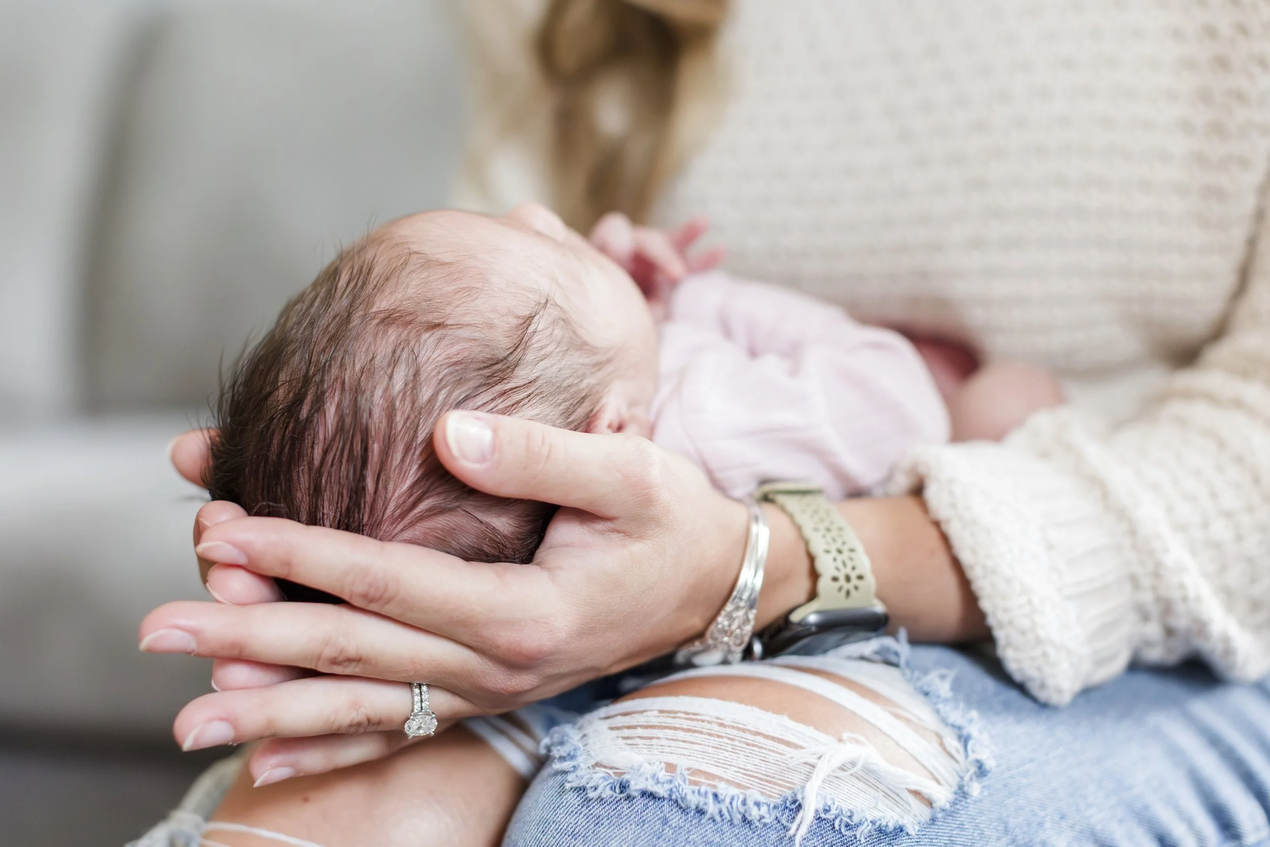 A woman holding a newborn baby close, with the baby's head resting in her hand. The woman is wearing a beige sweater and ripped jeans.