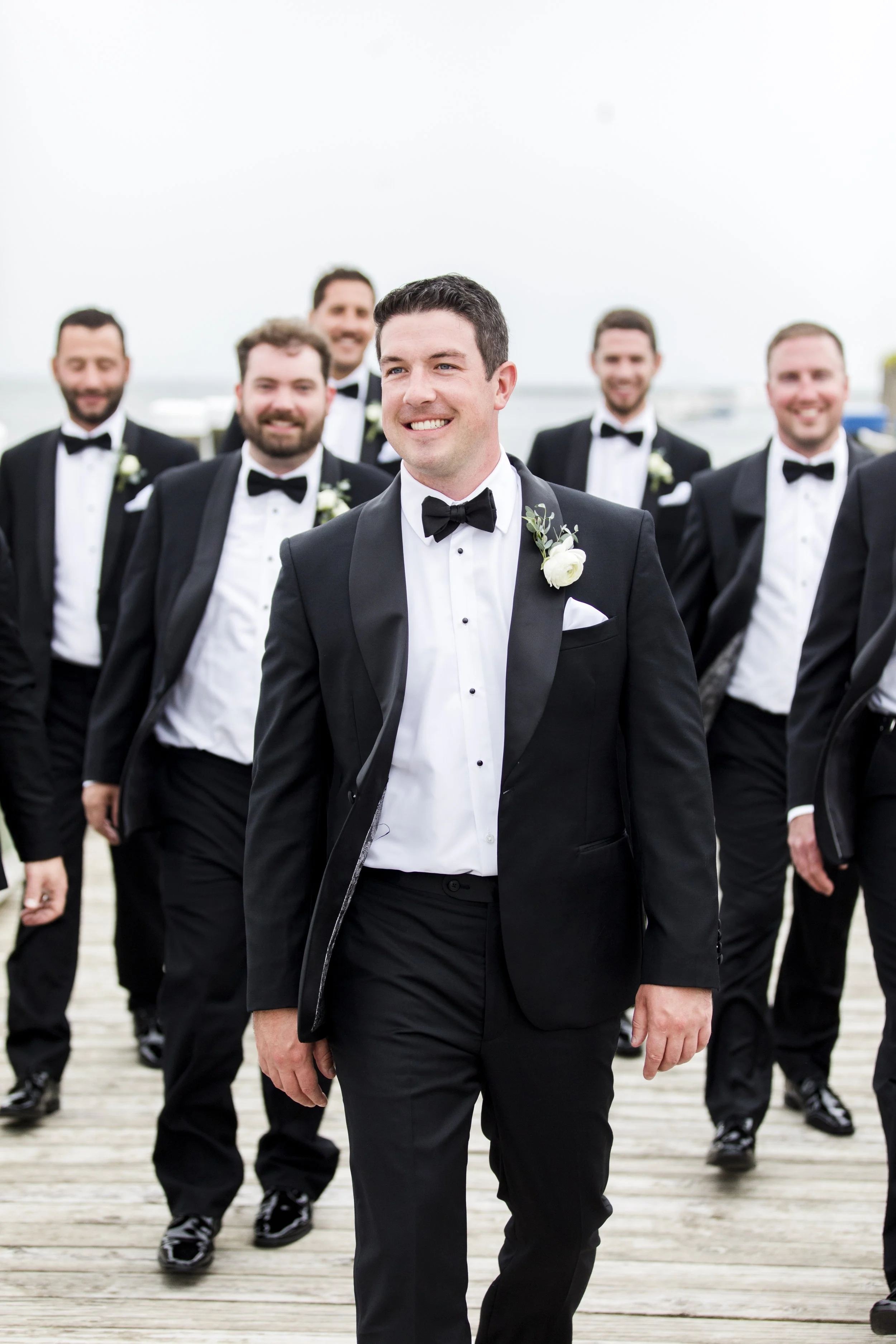 Group of men in tuxedos walking together on a wooden dock, with the groom in front smiling.