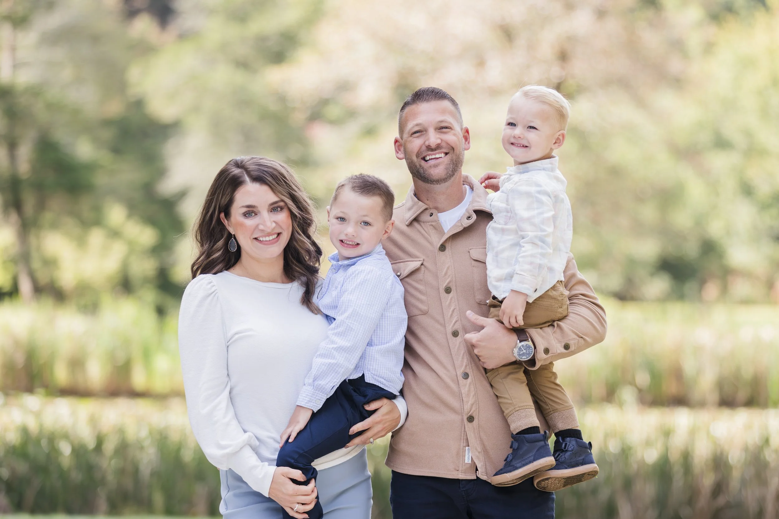 A family of four posing outdoors, with a woman and man holding two young boys in their arms, surrounded by trees and greenery.