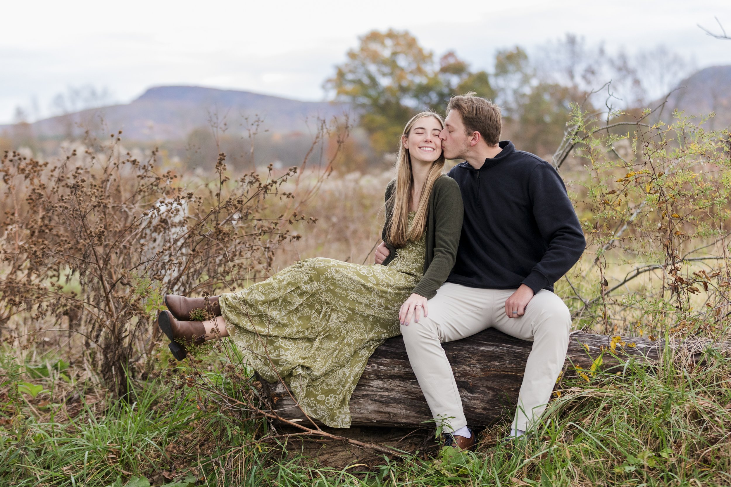 A young couple sitting on a log in a field, sharing a kiss, with fall foliage and mountains in the background.