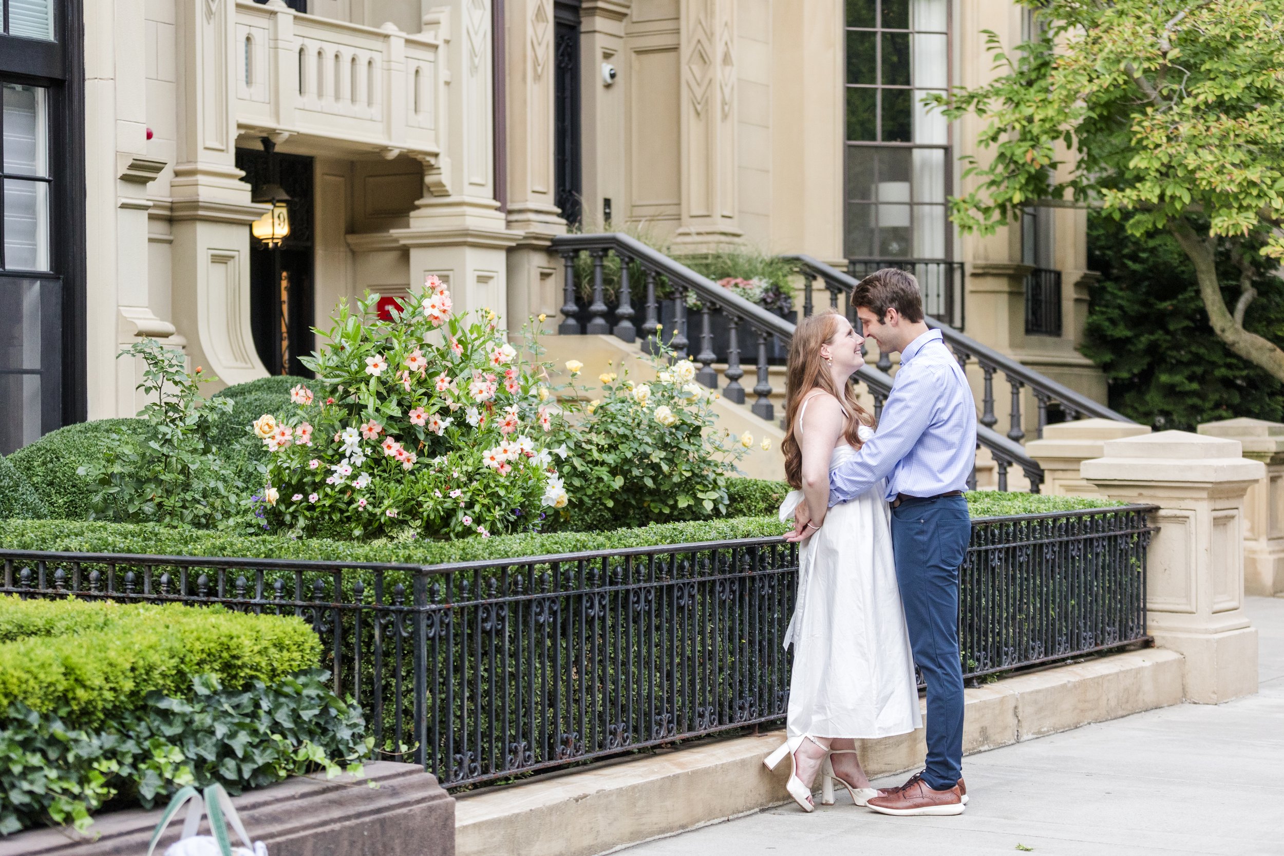 A couple is standing close together in front of a garden with blooming flowers and greenery, in front of a beige building with stairs and black railings, sharing an intimate moment.