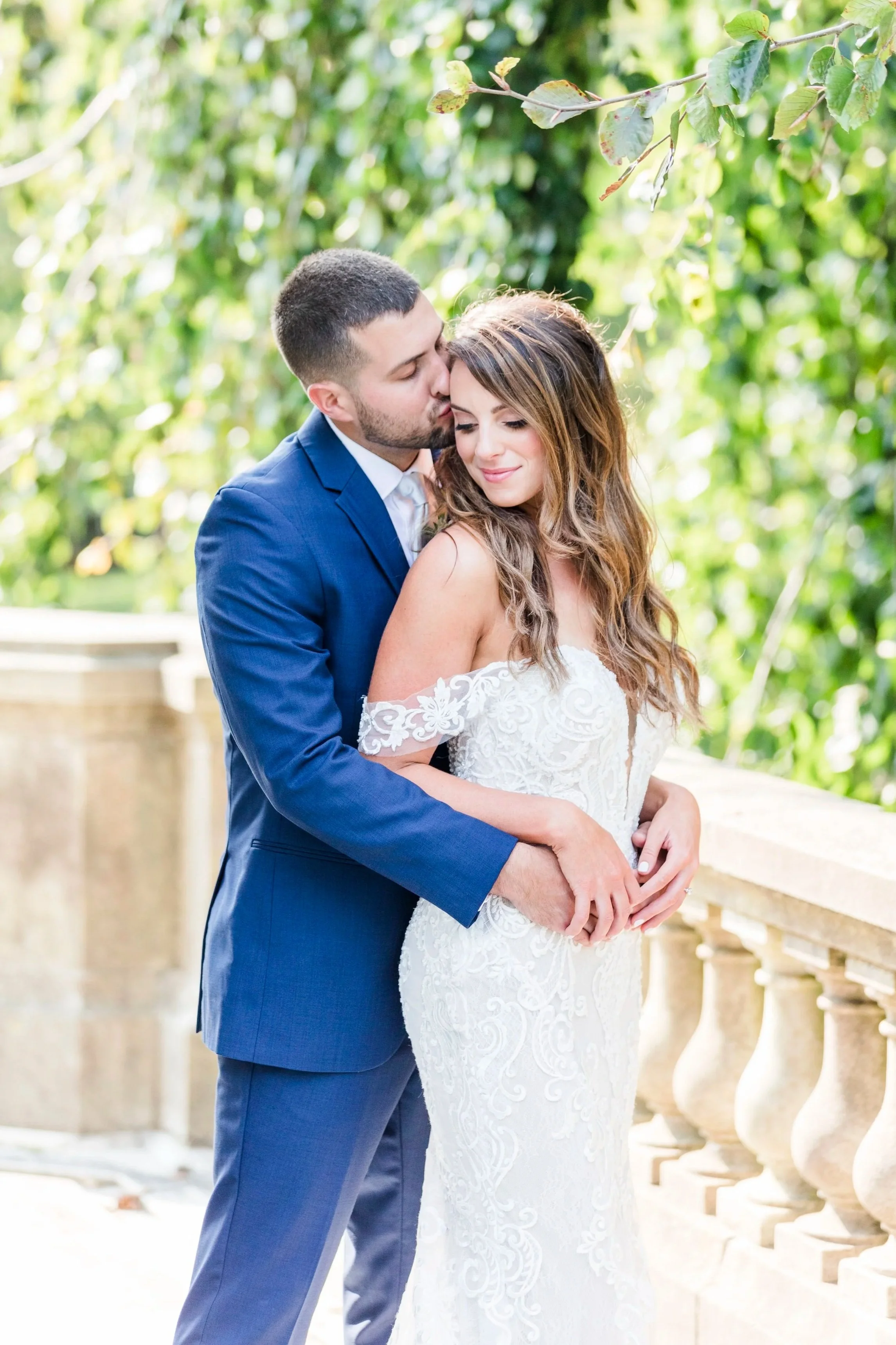 A couple on their wedding day, the groom in a blue suit kissing the bride on her forehead, outside with greenery in the background.