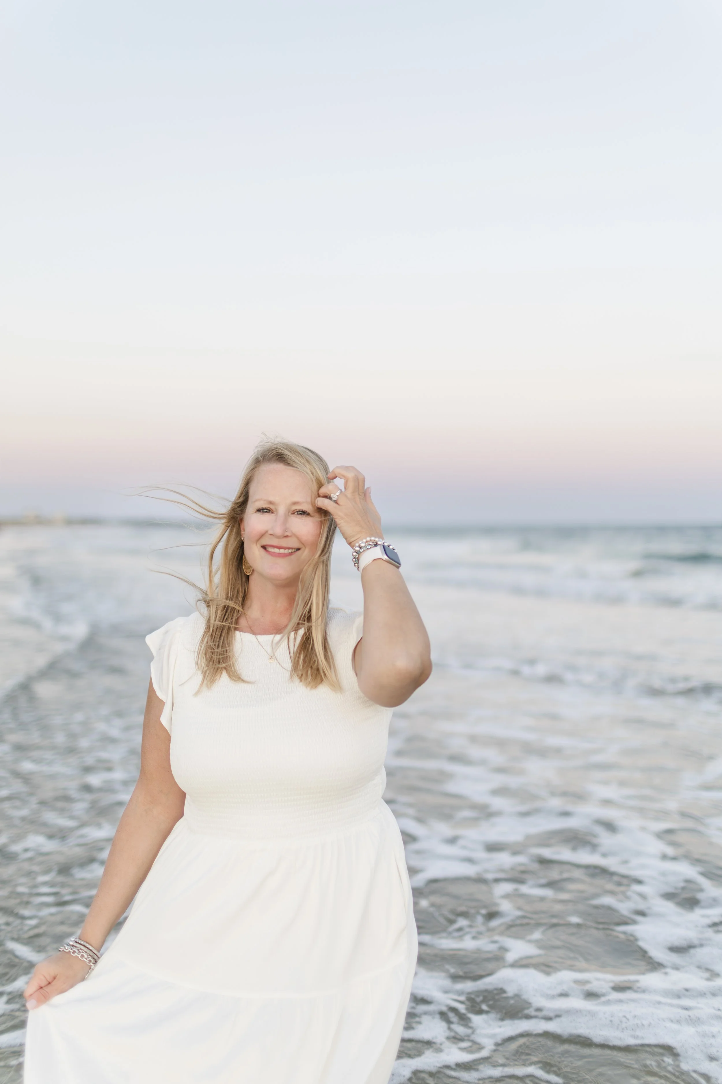 A woman in a white dress standing on the beach with waves at her feet, smiling at the camera during sunset.