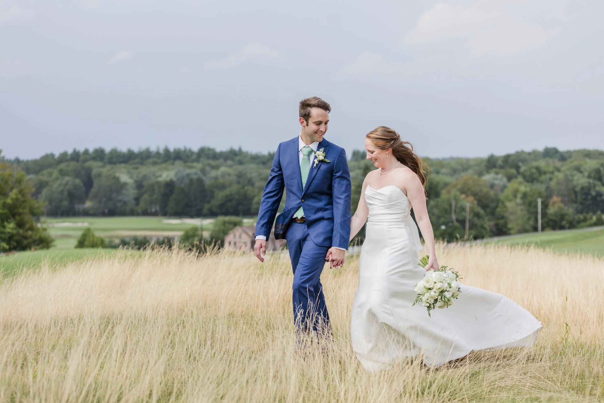 A bride and groom walking hand in hand through a grassy field on their wedding day, with a scenic landscape of trees and hills in the background.