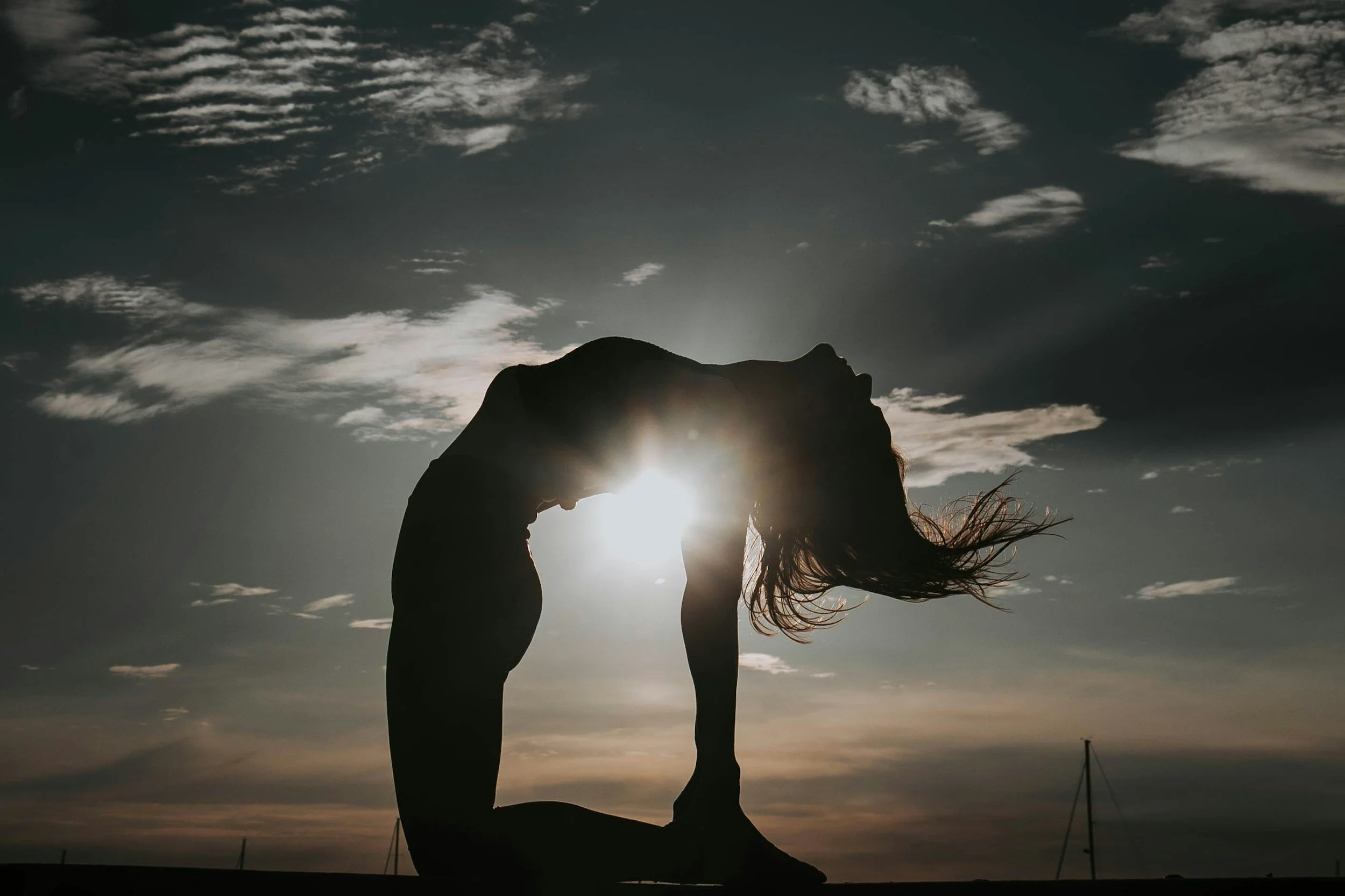 A silhouette of a woman doing a forward bend yoga pose outdoors at sunset, with the sun positioned behind her and her hair flowing in the wind.