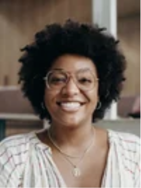 A woman with curly black hair, glasses, and a big smile, wearing a striped shirt and jewelry, inside a room