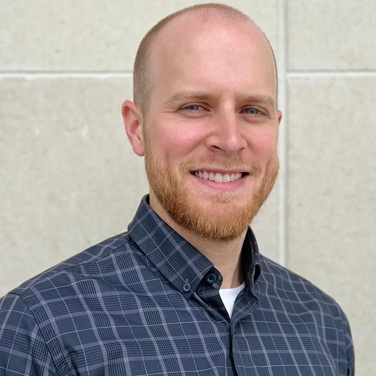 A smiling man with a beard and short hair wearing a blue checkered shirt standing in front of a beige wall.