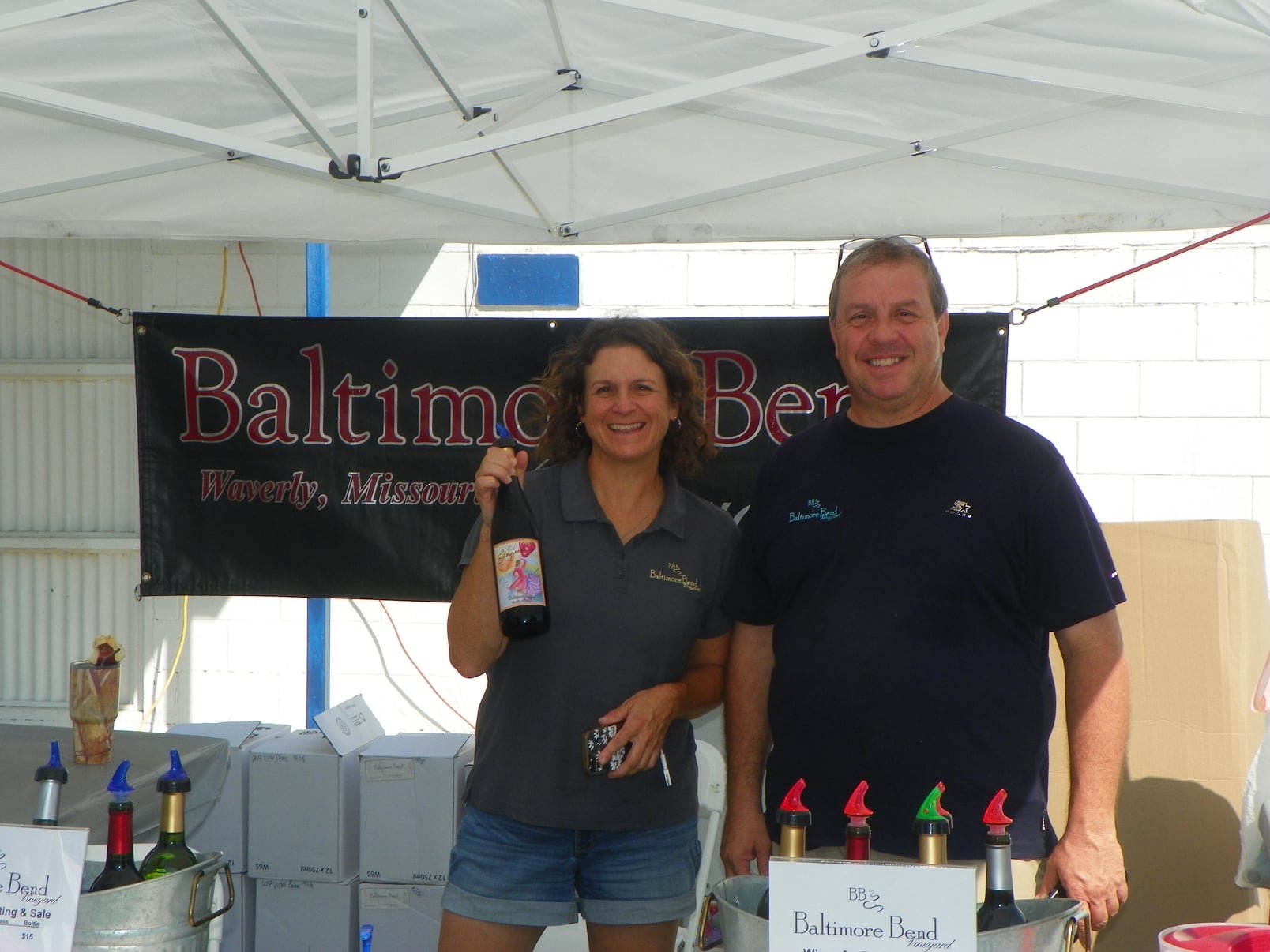 A woman and a man smiling at a Baltimore Bend winery booth, with the woman holding a bottle of wine. Behind them is a black banner with red and white text, and there are wine bottles and pouring spouts on the table.